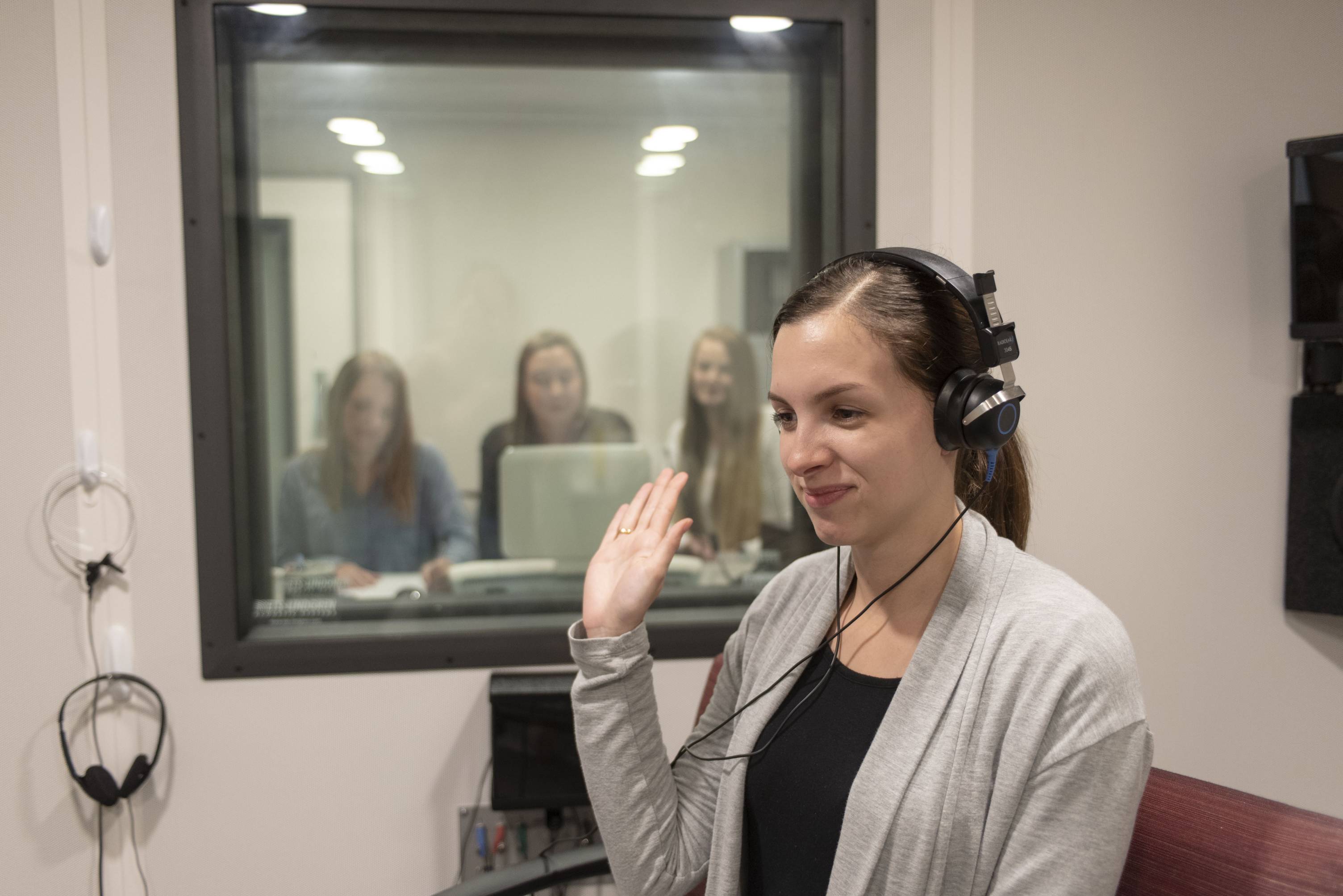 A young woman wearing headphones holds up her hand, faintly smiling. Behind her is a window to another room, where three other women observe and operate a computer.