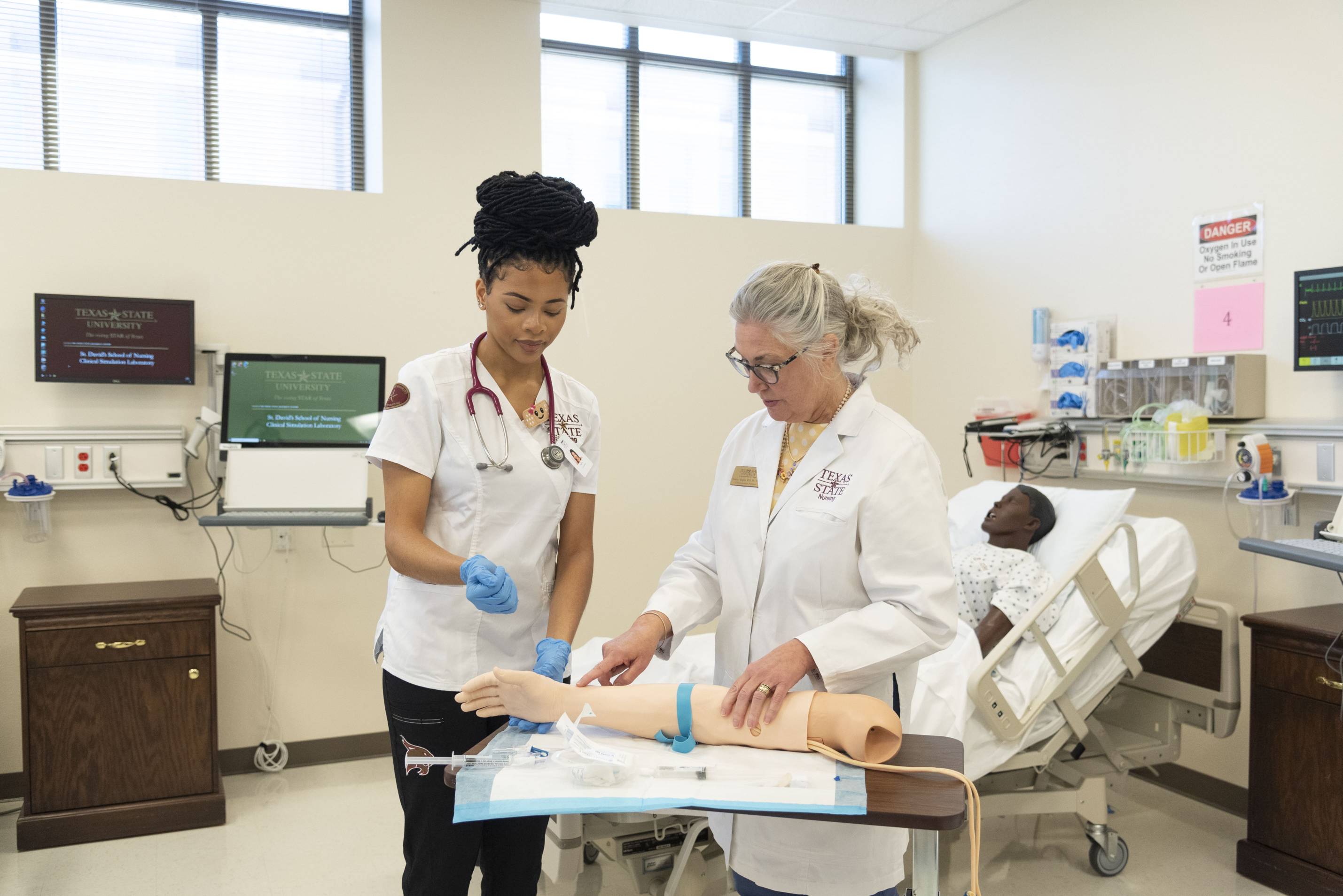 In a hospital setting an older woman wearing a labcoat gestures to a dummy patient arm. A younger woman wearing a labcoat, stethoscope, and blue gloves listens to her directions as she looks at the dummy.