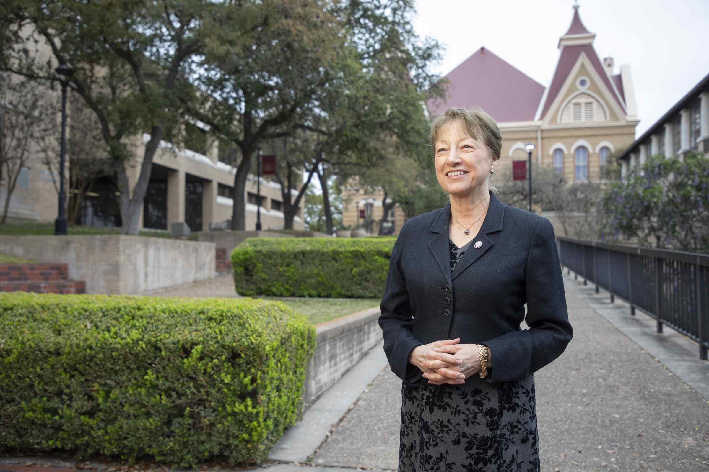 A woman wearing a black skirt suit clasps her hands and looks into the distance, her eyes wistful. Behind her can be seen a building and manicured hedges.