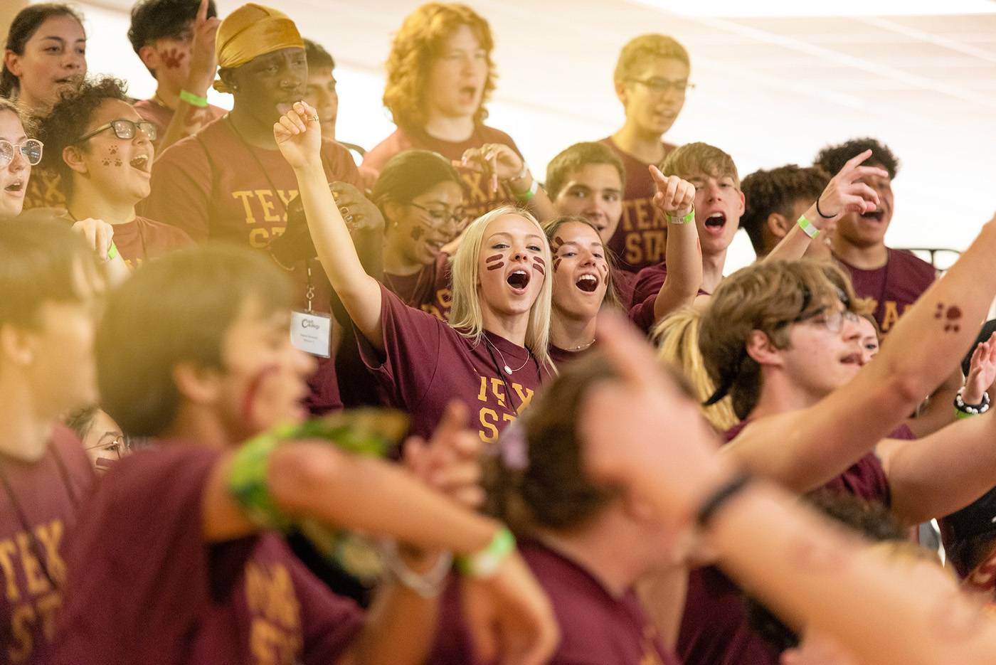 Close up shot with a lens flare of a group of cheering young people. They all wear maroon school spirit T-shirts and some have face paint.