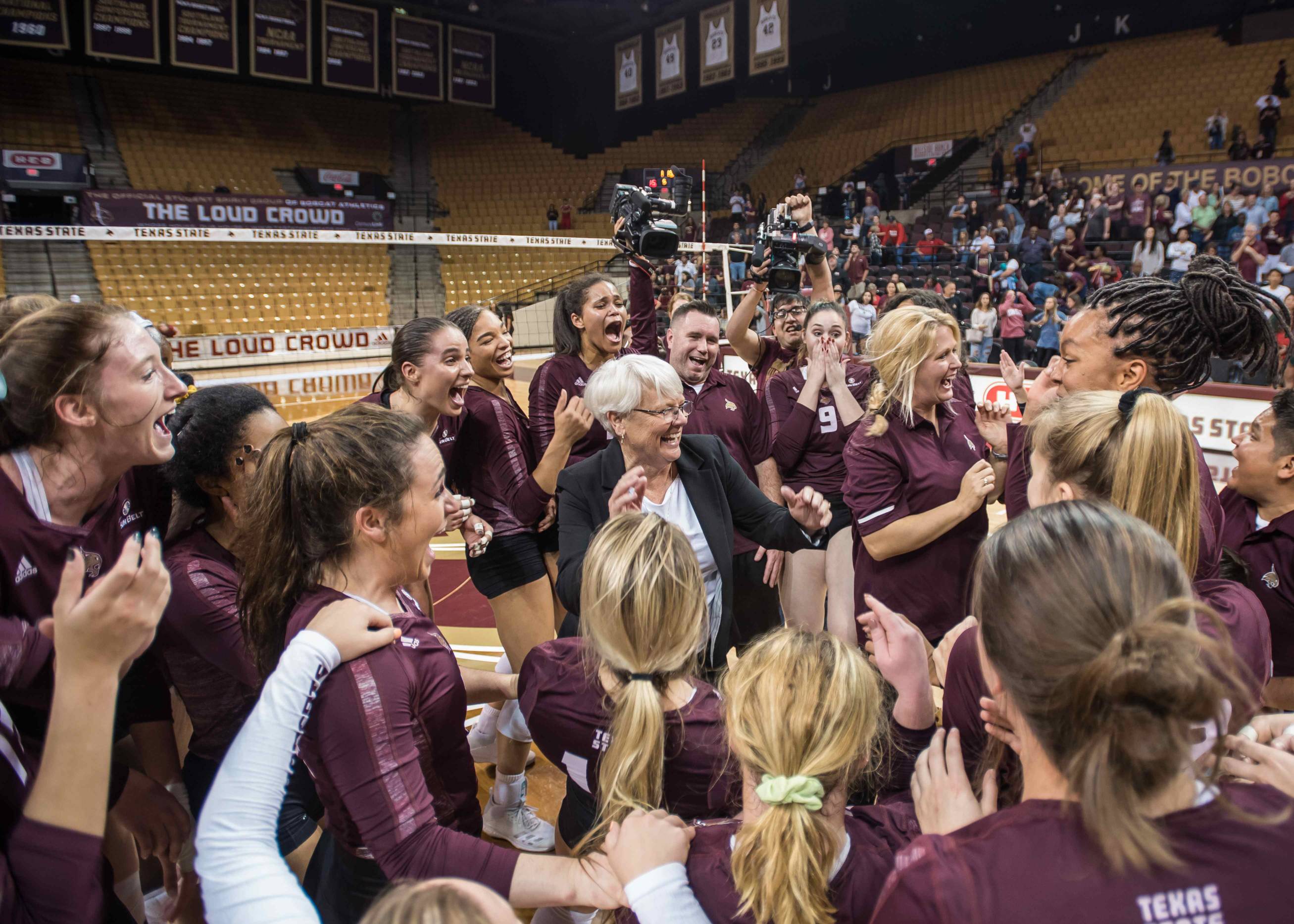 An older woman with a shock of white hair is surrounded by emotive young women in maroon uniforms.