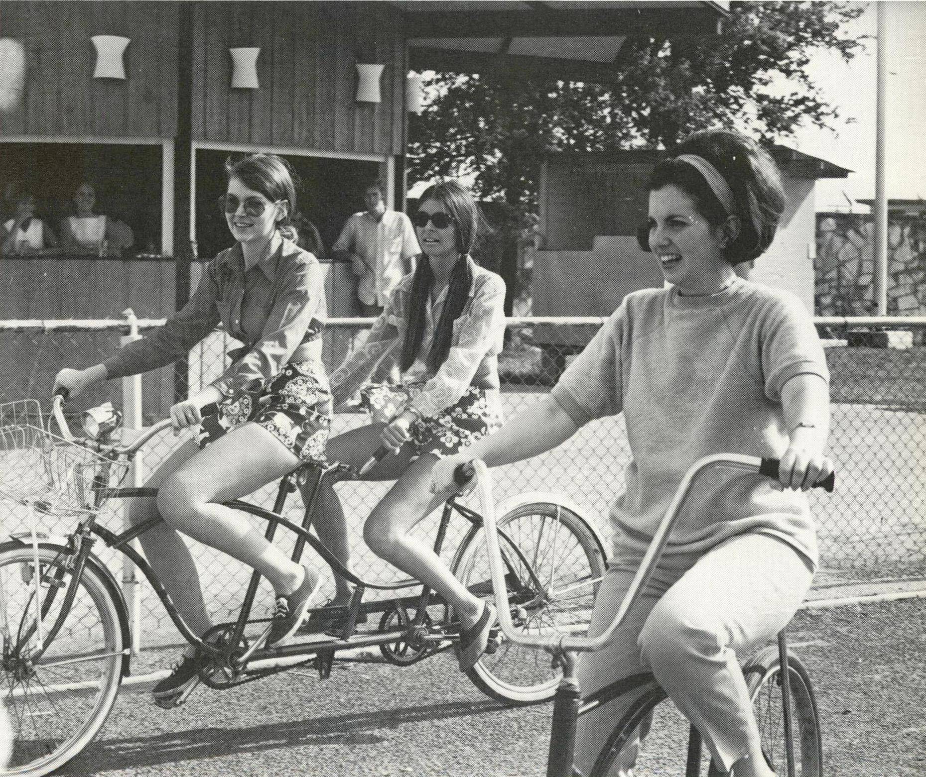 Candid black and white photo of three girls laughing and smiling on a sunny day in the 1970s. Two of the girls ride a tandem bike down a road and a third, with a teased bob hairstyle, rides a single bike.