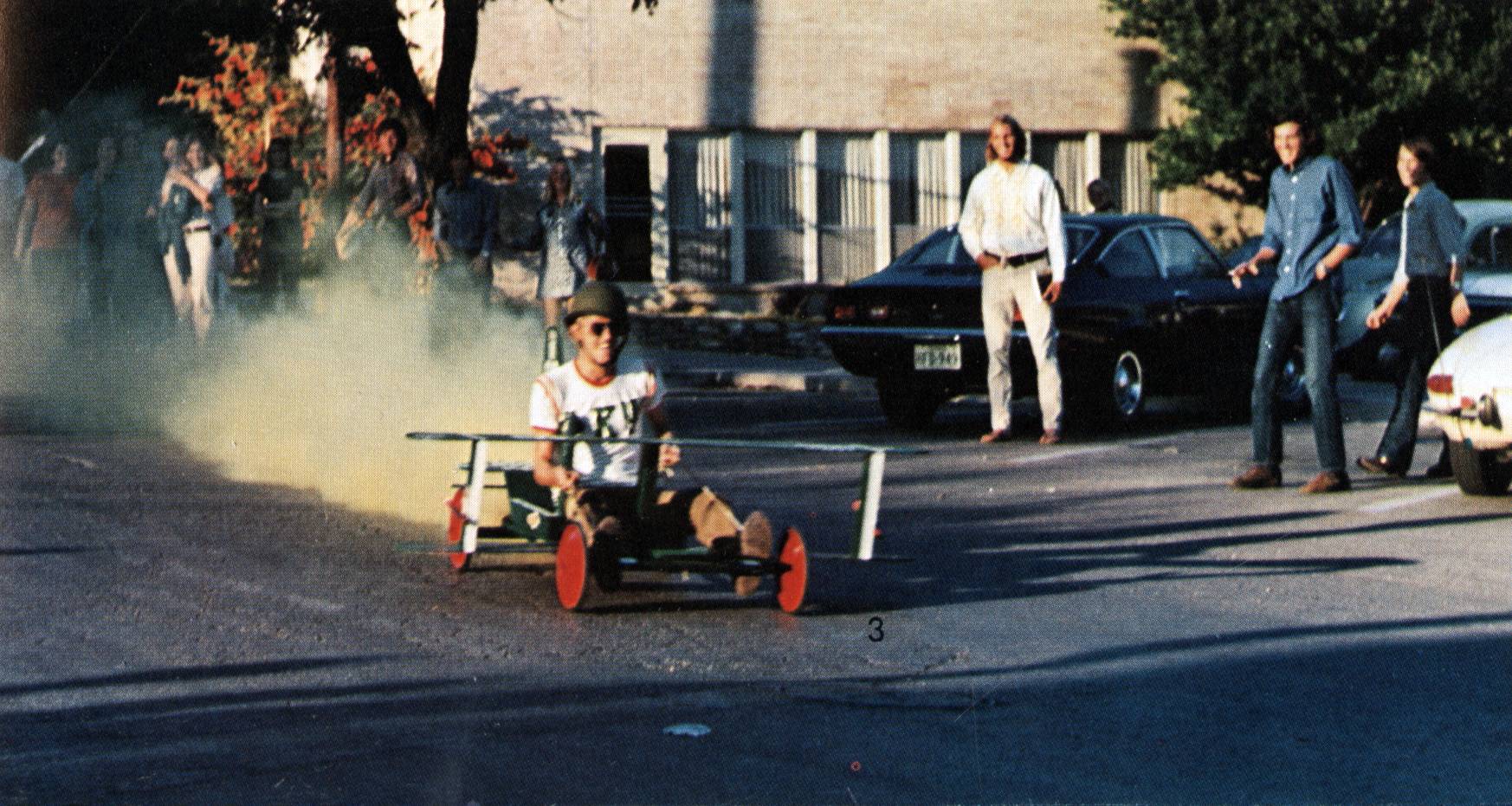 Color photo of a young man driving through a parking lot in what appears to be a homemade derby car. To his right, three other men survey the derby car's progress. Behind him and to the left are a group of people cheering.