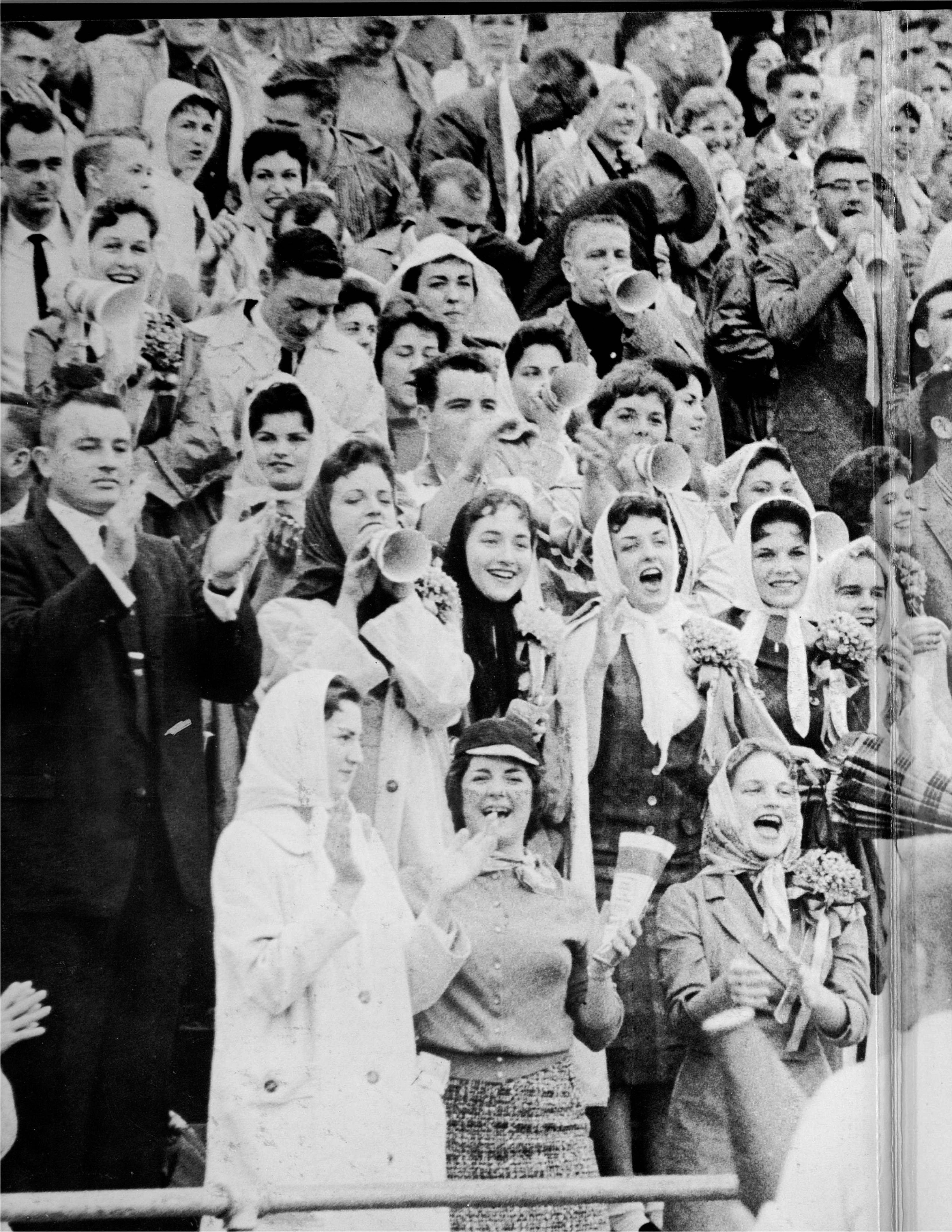 Black and white photo of young men and women cheering and standing on bleachers. Many of them wear sweaters or pea coats and the women wear hats or scarves tied around their heads.