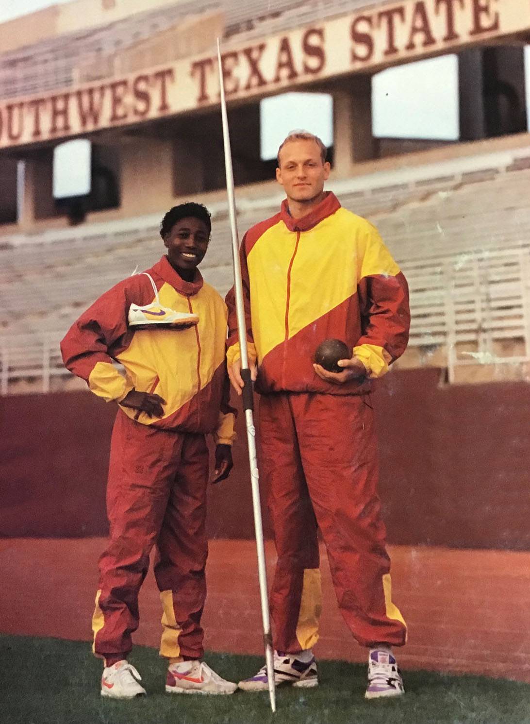 A man and a woman smile and pose for the camera in bright yellow and red tracksuits. The woman, left, has a pair of running shoes slung over her shoulder. The man holds a pole in one hand and a ball for shot put in the other.