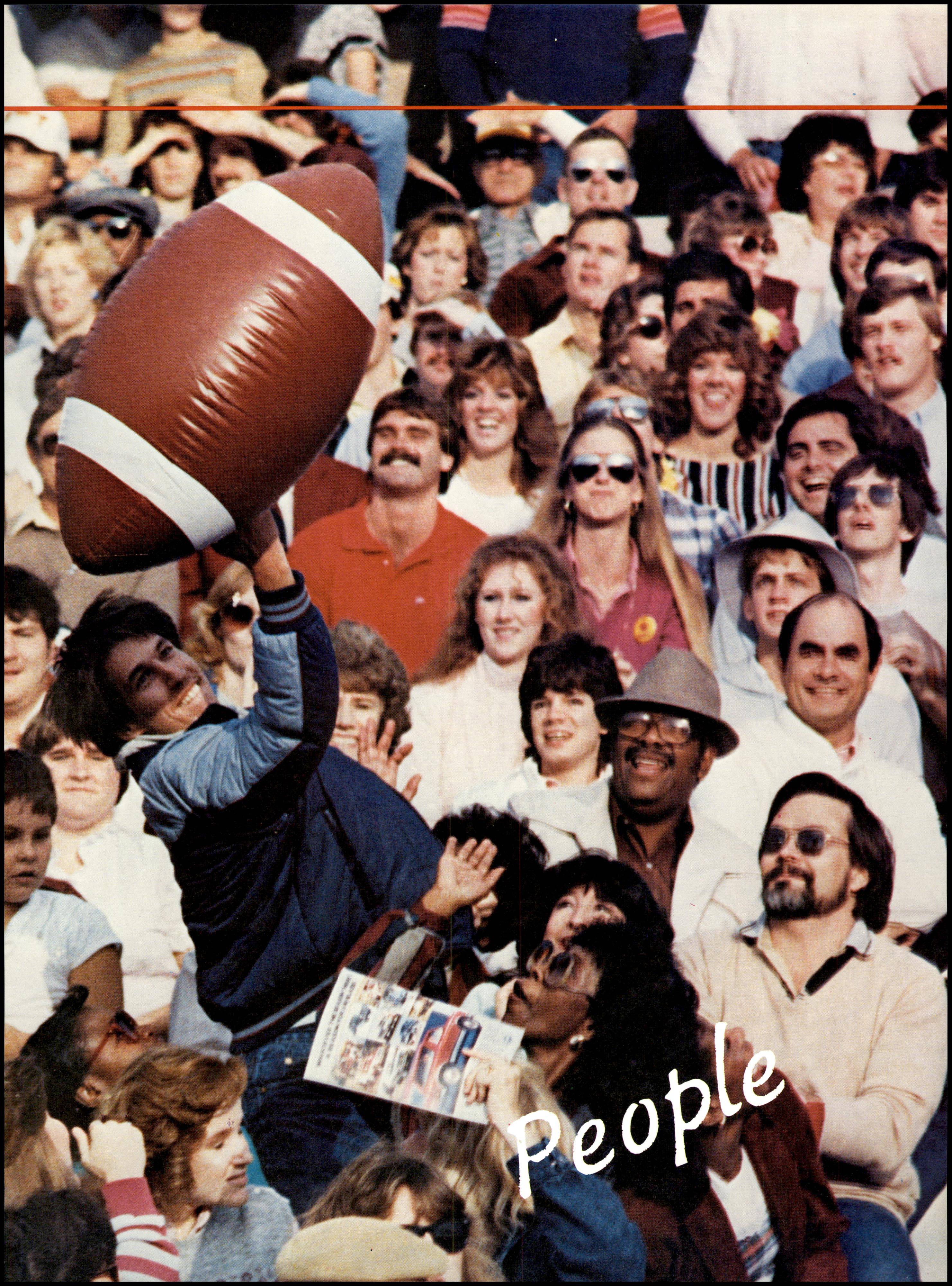 A young man jumps up to bat a large inflatable football toy. Behind him are seated football fans in the stands, their outfits and hair indicating the 1980s.