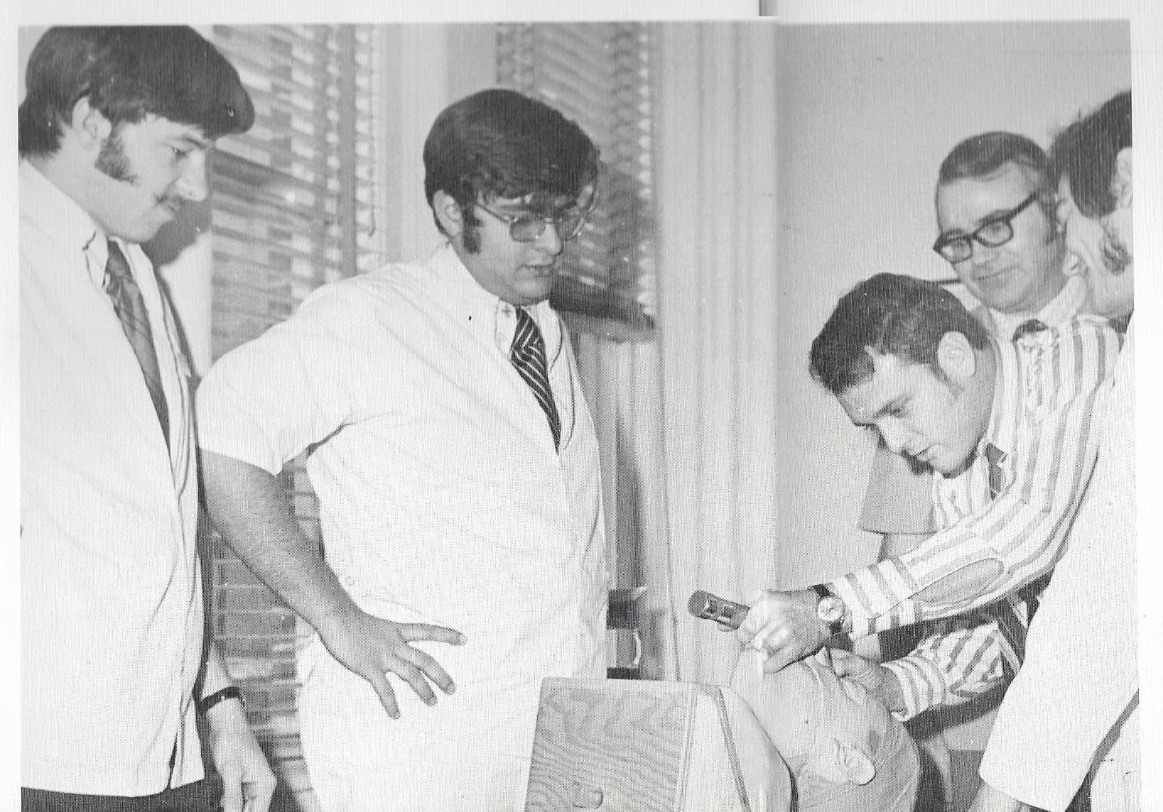 Black and white photo of a group of five men performing a mock procedure on a dummy patient. Several of the men wear labcoats and sport sideburns and thick, black glasses.