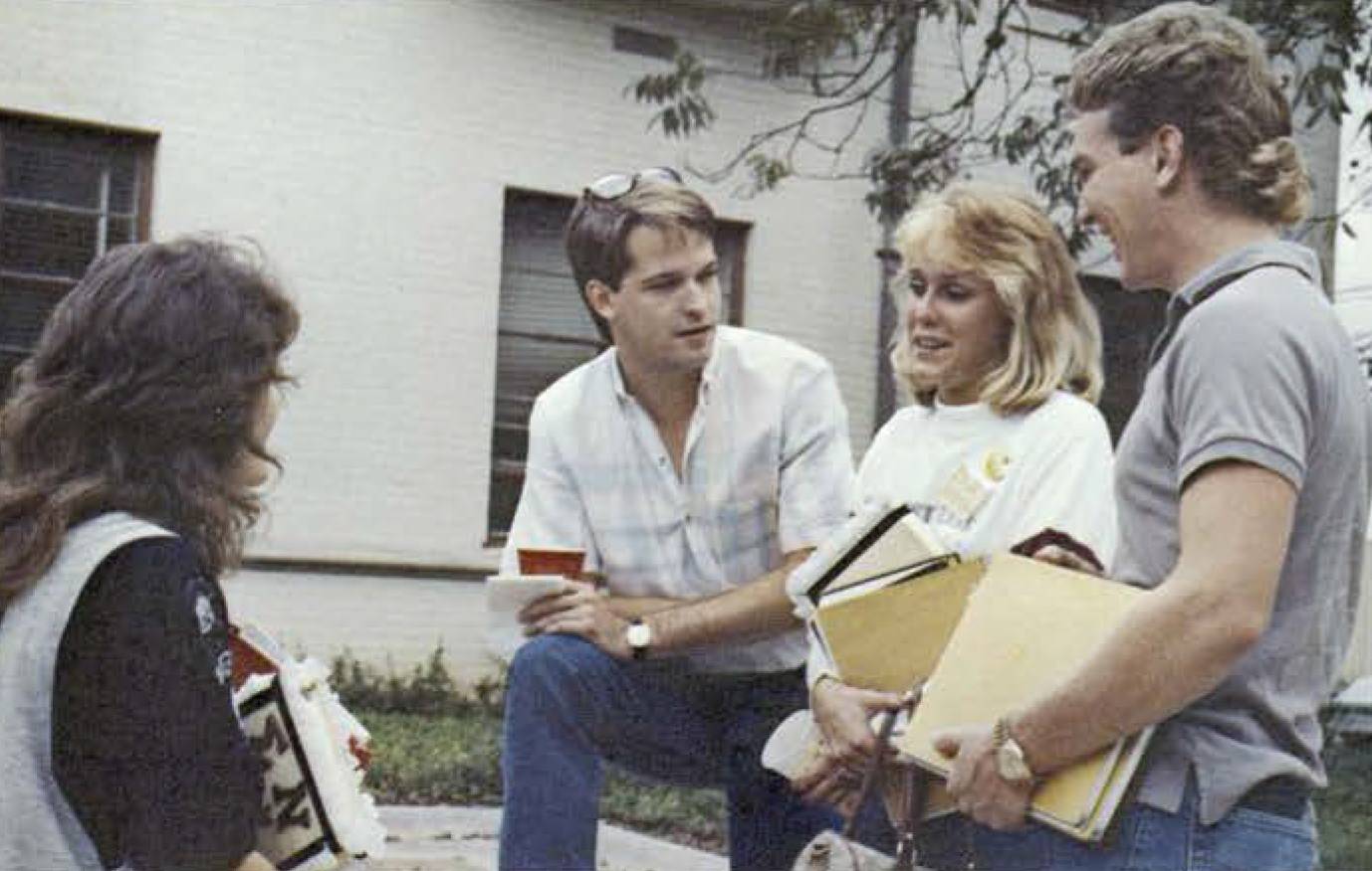 Four young men and women, all with feathered hair, stand in a loose circle and chat. Three of them hold stacks of books and the fourth holds a red cup.