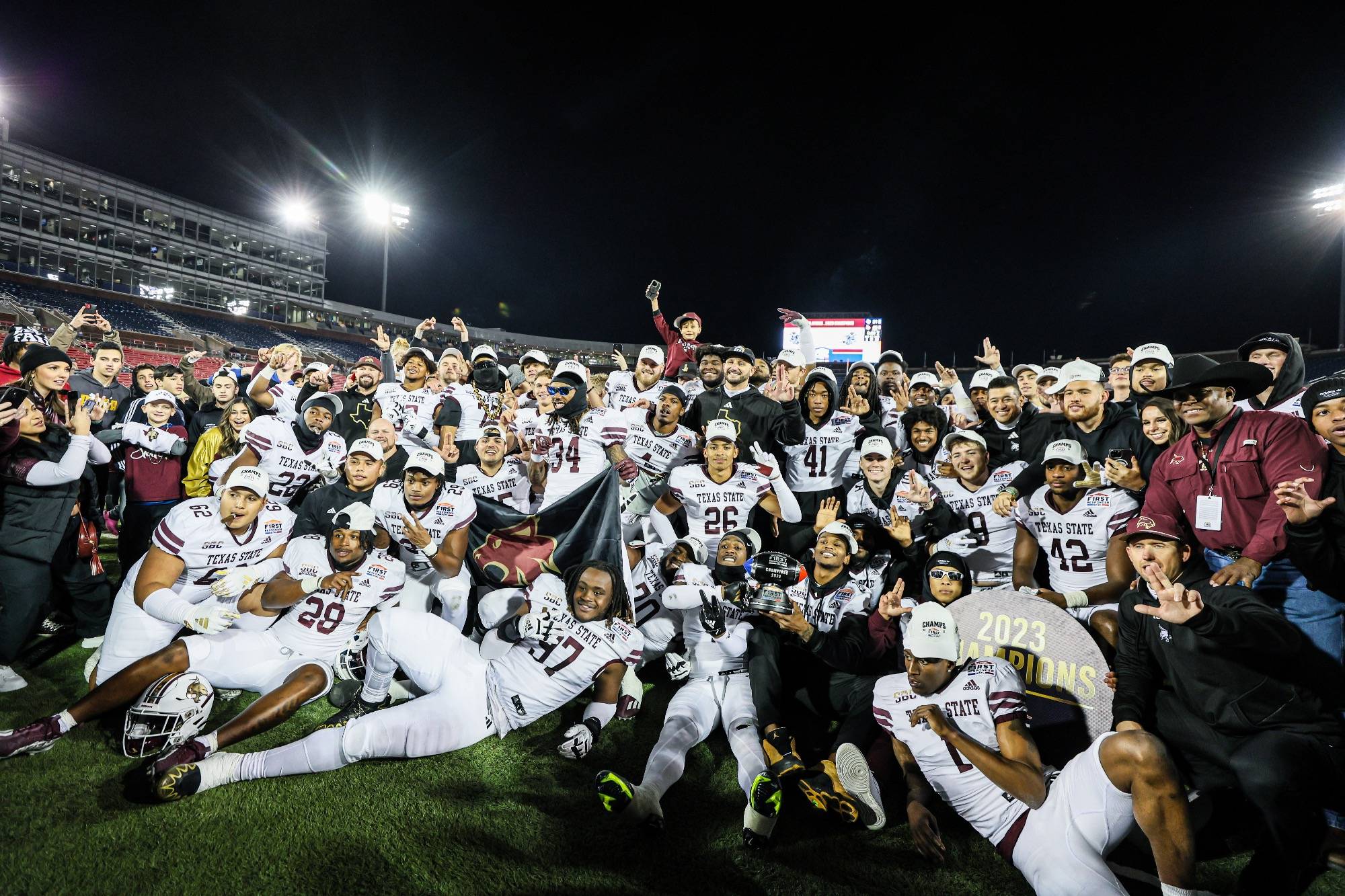 A group of young men in football uniforms pose confidently on a dark football field, holding a sign that reads "2023 champions".