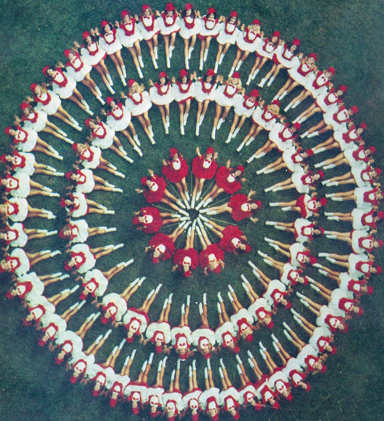 Aerial shot of young women wearing bright red and white dresses and hats. They're sitting in the three concentric circles on grass, looking up at the camera.