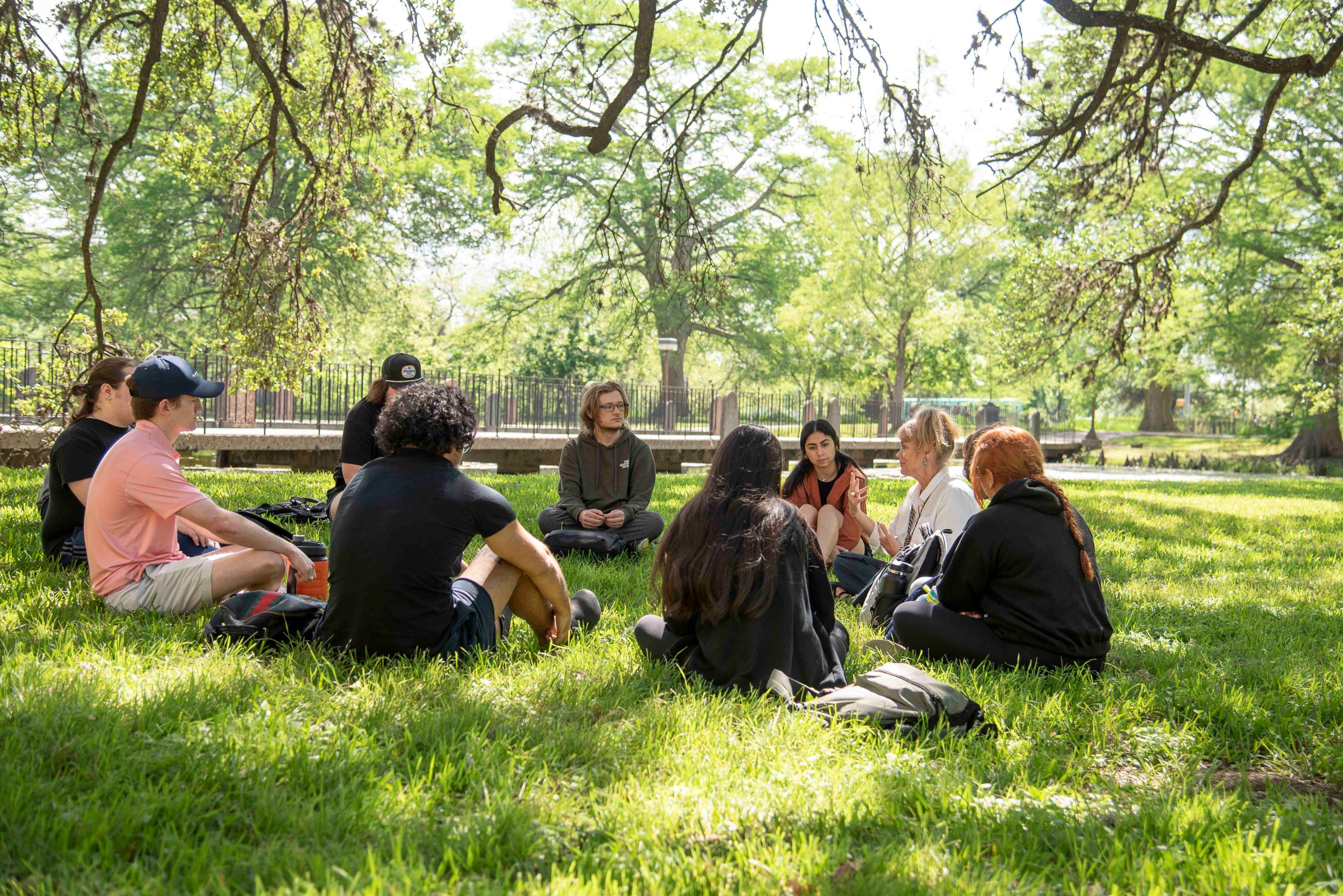 A group of young people sit on the ground in a circle, looking intently towards an older woman. The ground is grassy and sunlight shines through the tree branches overhead.