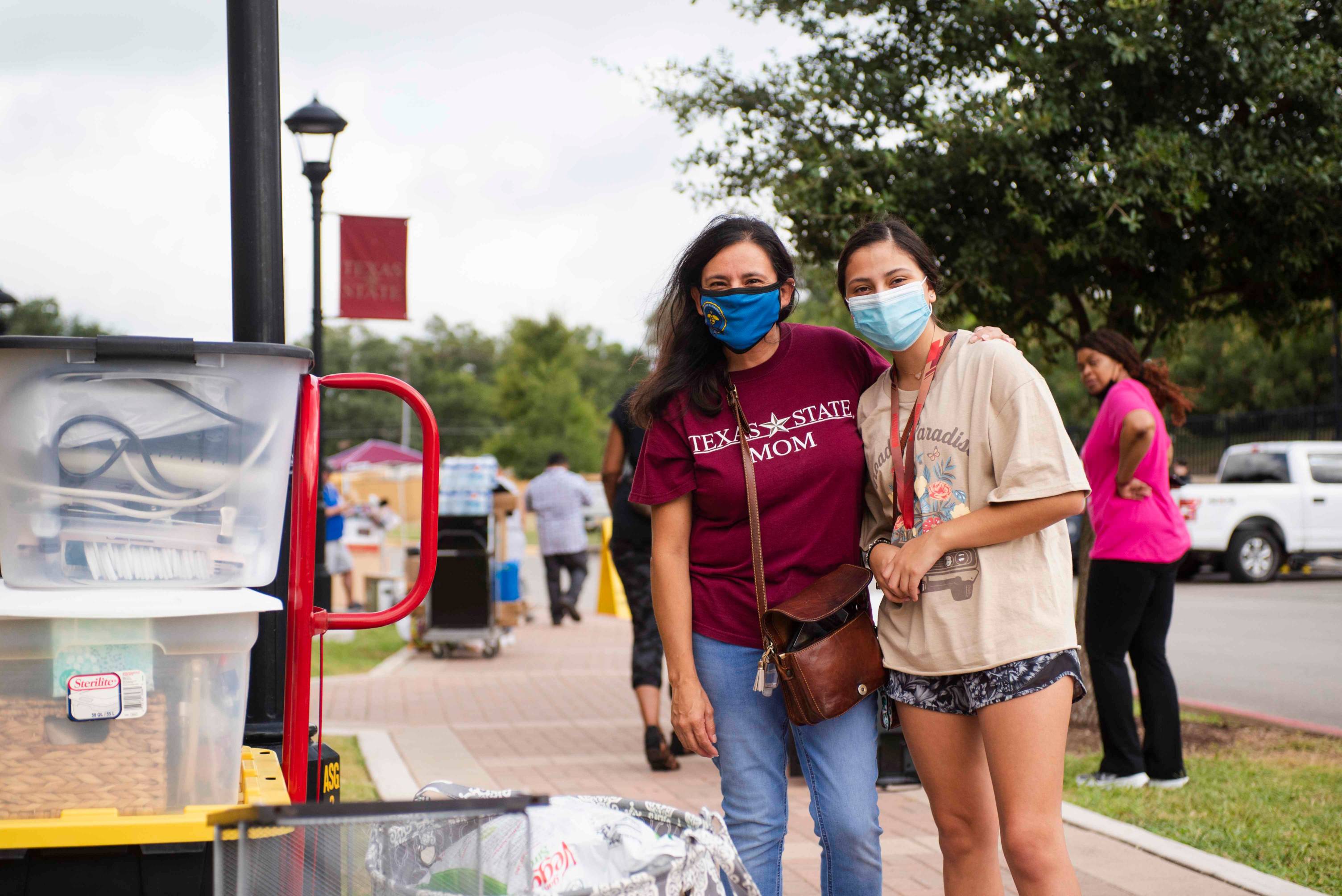 A mother and daughter, both wearing face masks, stand together on a sidewalk in front of storage tubs of one's belongings.