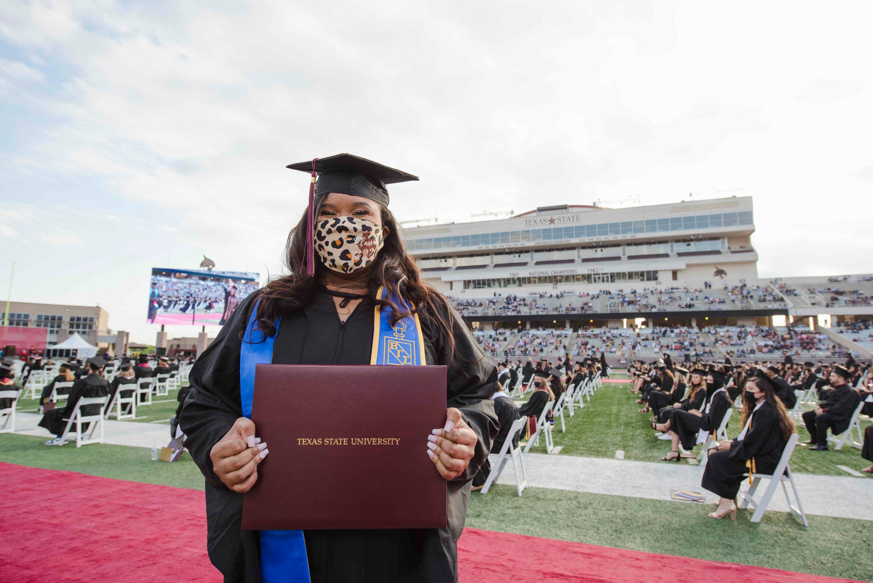 A girl in a graduation cap and gown and cheetah print face mask holds her diploma on a football field. Rows of seated graduates can be seen behind her.