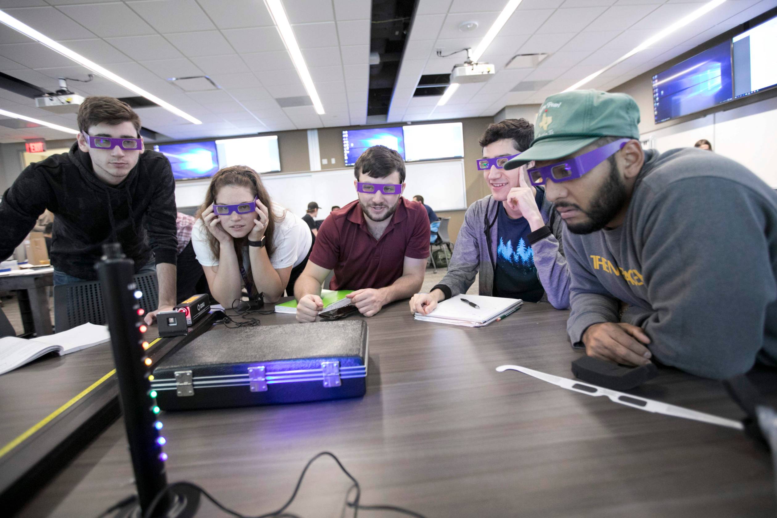 Five students sit at a table. They are all wearing purple, specialty glasses and looking at a device emitting colors across the light spectrum.