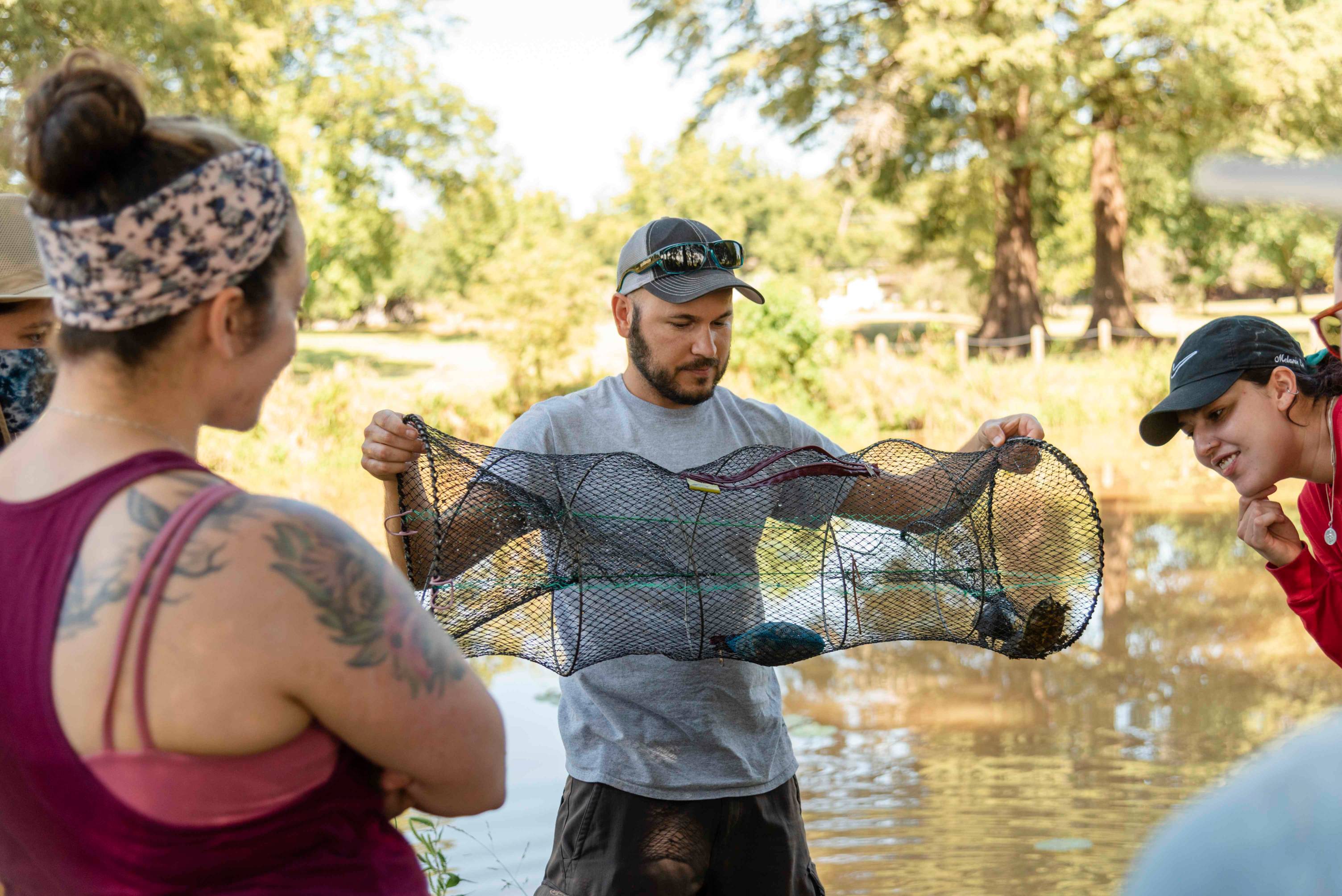 A man in casual dress with sunglasses on top of his cap holds a mesh tube with several turtles inside while several people survey. In the background, a river can be seen.