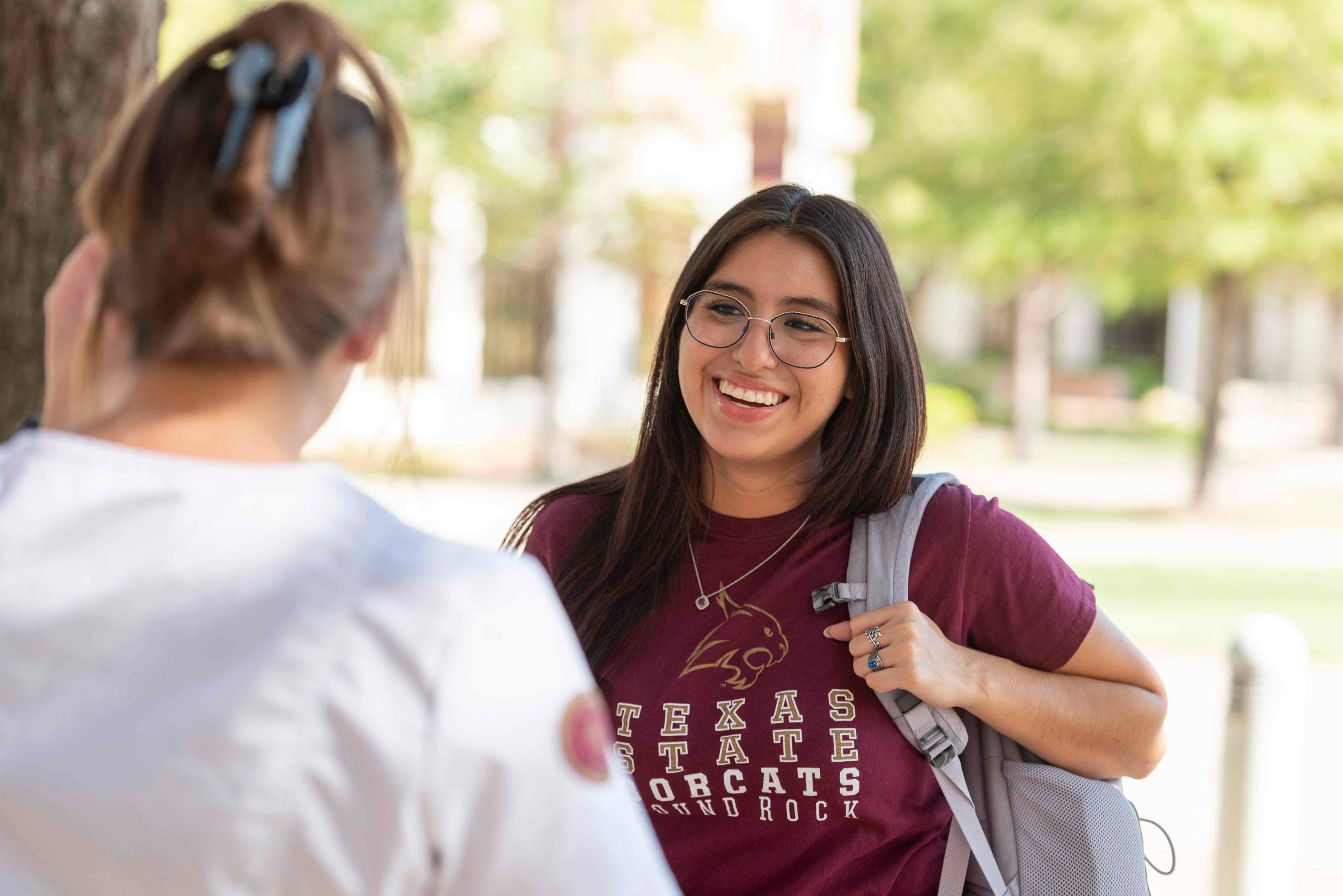 A photo of the view over a woman's shoulder while she talks to a young woman. The young woman is smiling and wearing a maroon university T-shirt and has a backpack slung over one shoulder.