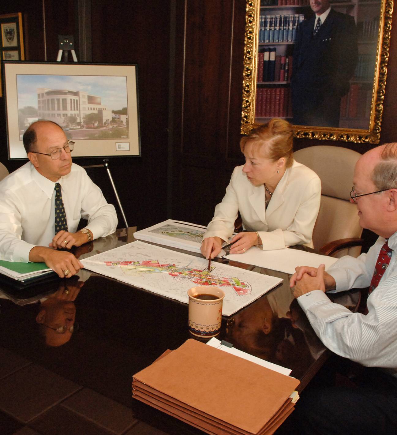 A woman in a white blazer sits at a glossy table, pointing toward a map. Two men in button down shirts and ties observe. On the table is a cup of coffee and a stack of papers. Behind them is an ornate, gold-framed painting and a drawing of a building sitting on an easel.