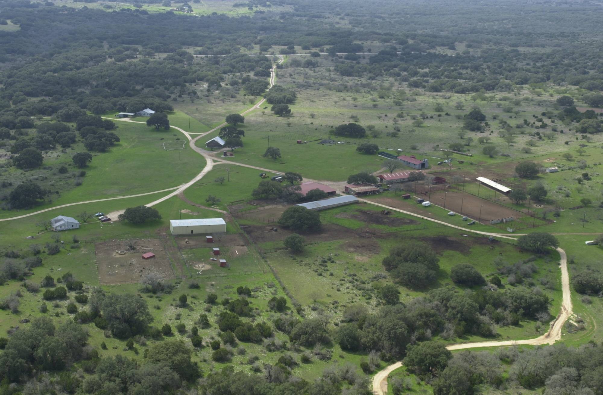 Aerial photograph of a pastoral scene. The land is grassy and dark green trees dot the landscape. A dirt road snakes up and around the property.