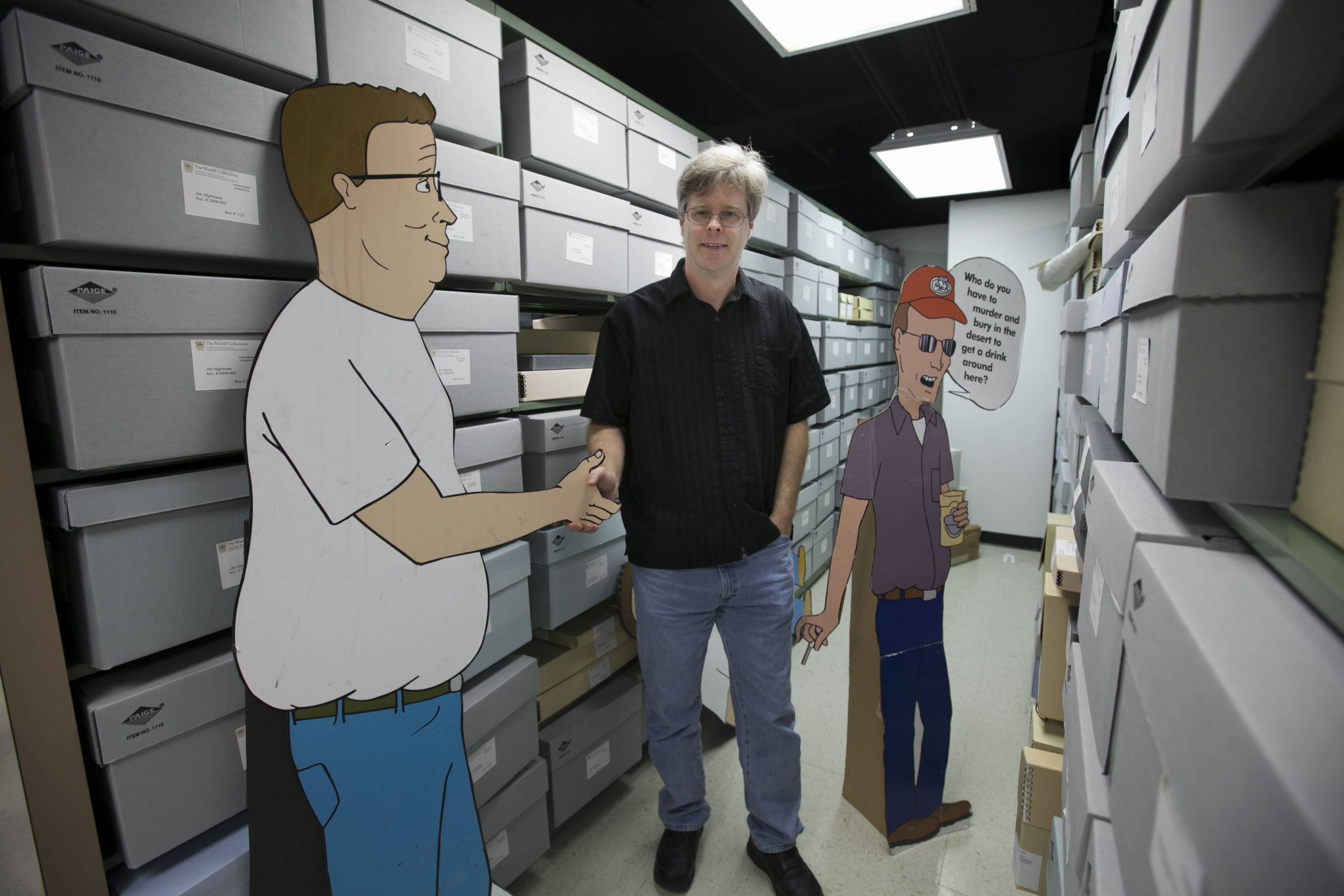 Bespectacled man in a polo shirt standing in an archive room with shelves and rows of boxes. The man is "shaking hands" with a cardboard cutout of Hank Hill, a cartoon character. Another character from the same cartoon, Dale Gribble, stands behind the man, with a speech bubble that reads: "Who do you have to murder and bury in the desert to get a drink around here?"