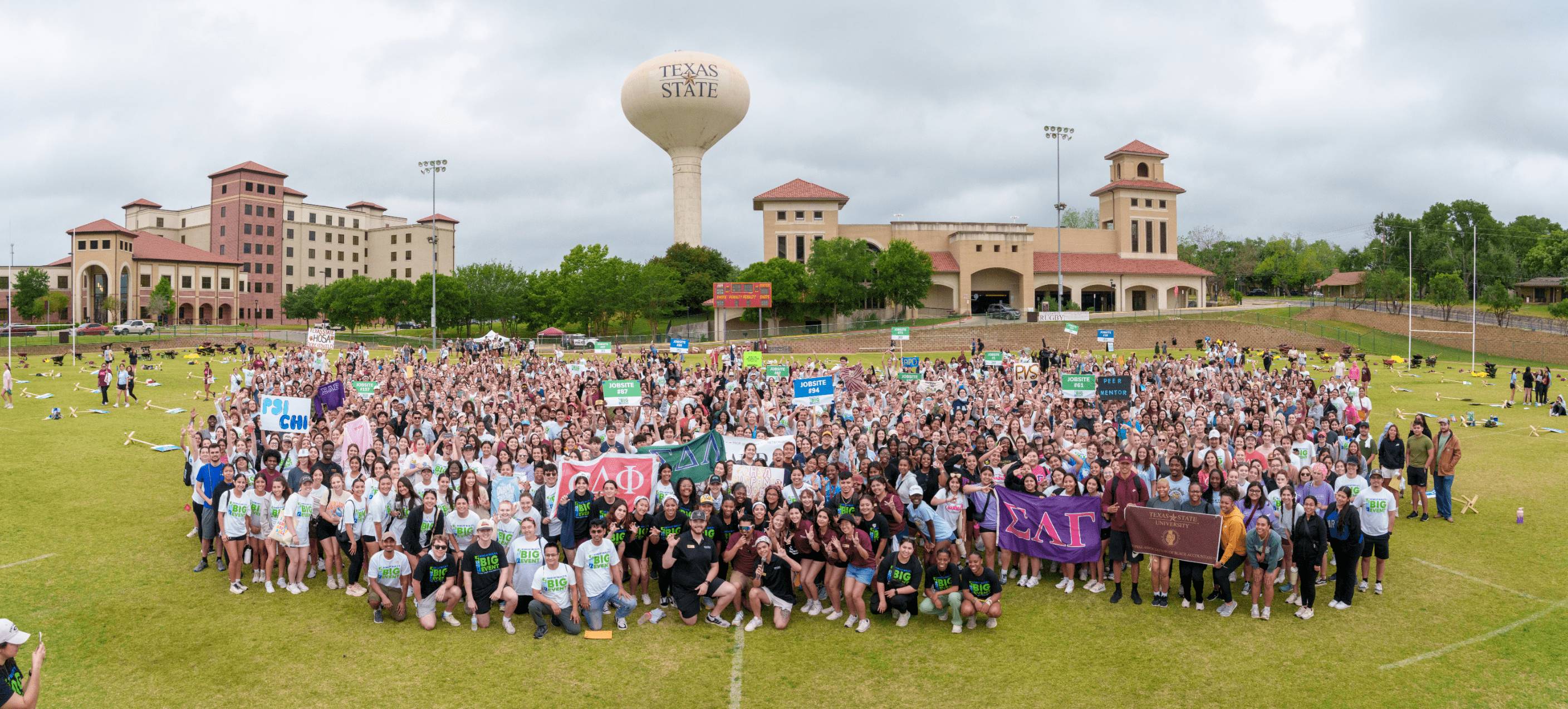 A crowd of several hundred people pose on a lawn, campus buildings and water tower behind them. Many of them clustered in matching outfits, holding aloft signs denoting Greek letters.