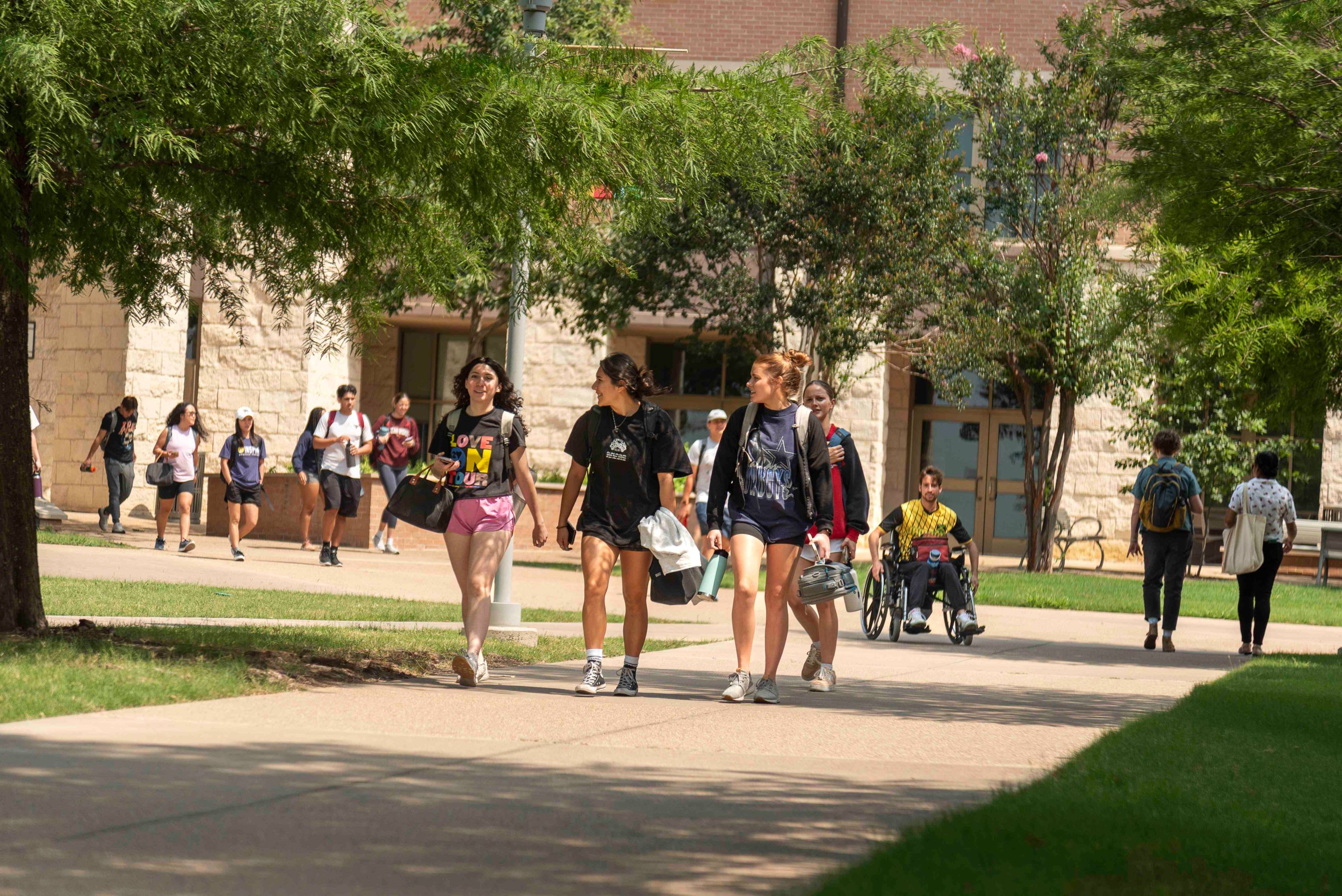 A group of young women chat as they walk down the sidewalk of a sunny college campus. Behind them is the impression of movement as students amble along.