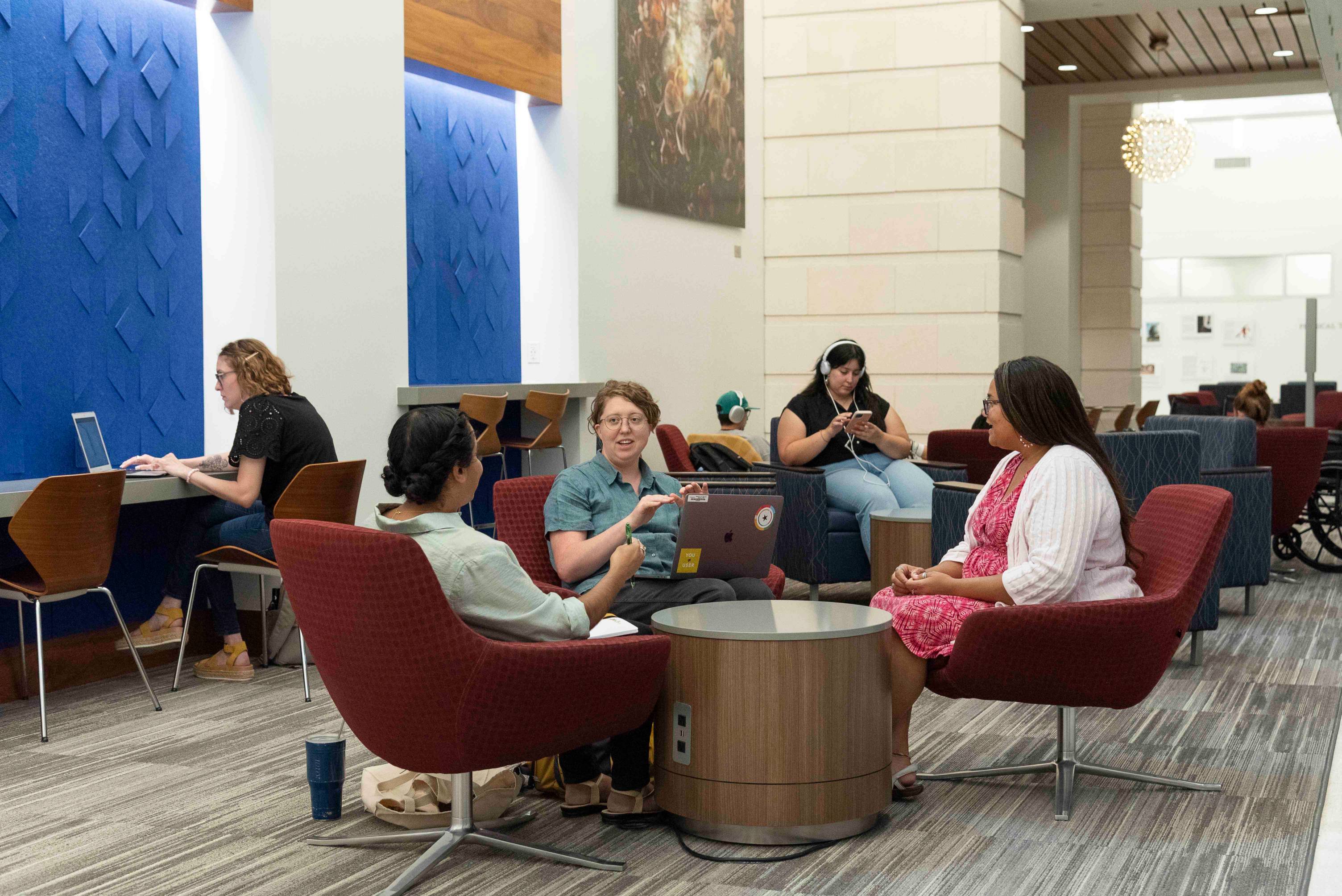 A group of young women sit in swivel chairs in a circle. They are engaged in conversation and one of them has a laptop on her lap. Behind them are various students engaged in study or looking at their devices.