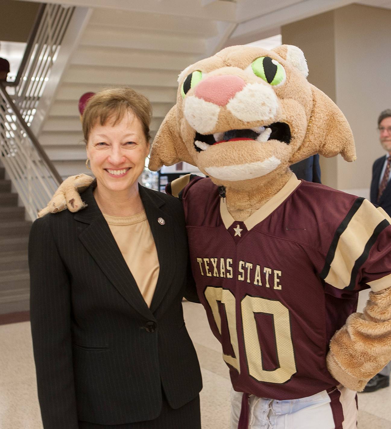 A woman wearing a blazer smiles, her arm wrapped around a person in a bobcat mascot costume. The costume is emblazoned with "Texas State".