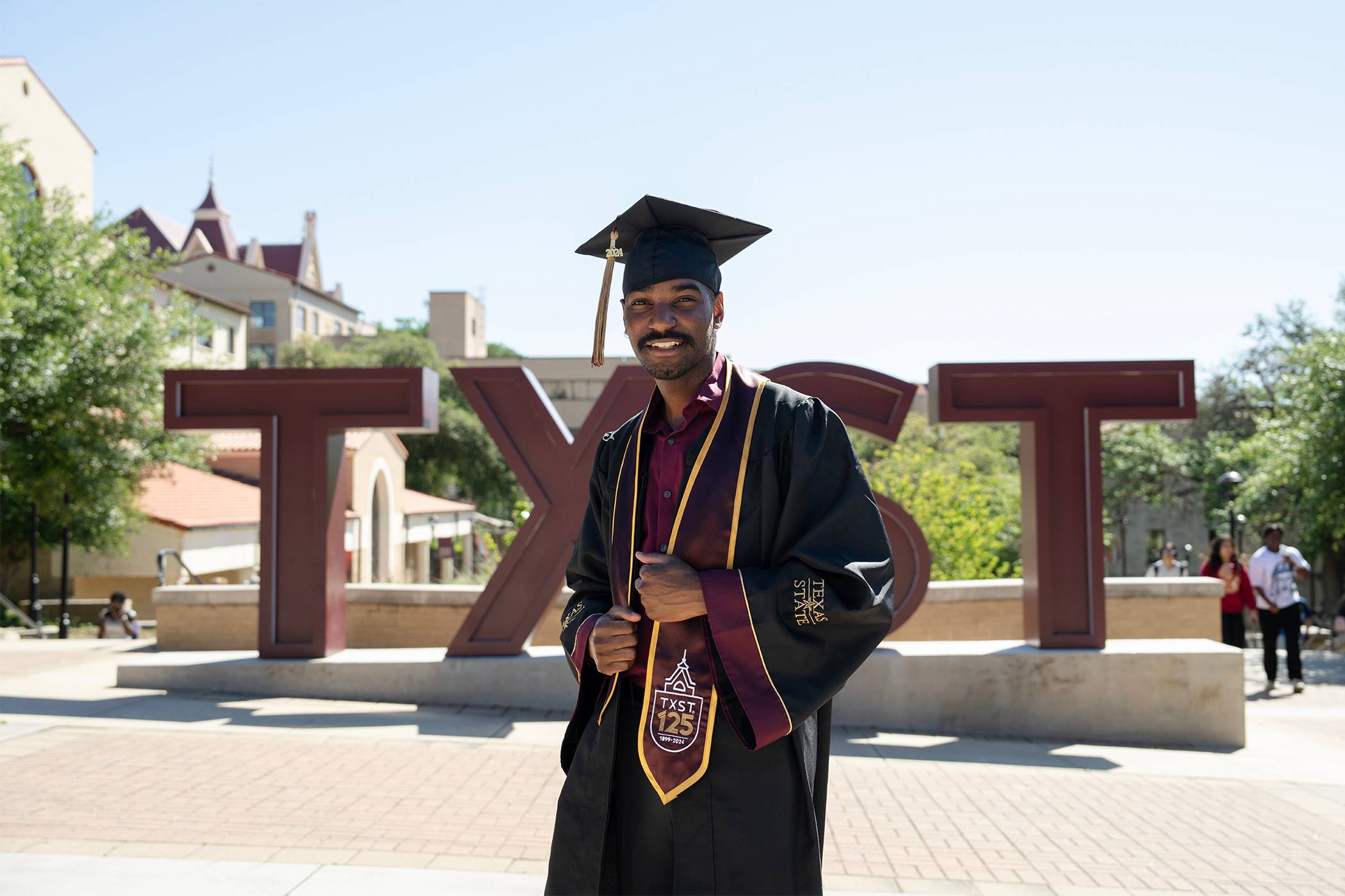 A proud young man dressed in his graduation regalia smiles for the camera as he adjusts his robes and stole. He's standing outside in front of the TXST statue on a bright and clear day, Old Main's distinct towers can be seen peeking out from behind the trees and buildings in the background.