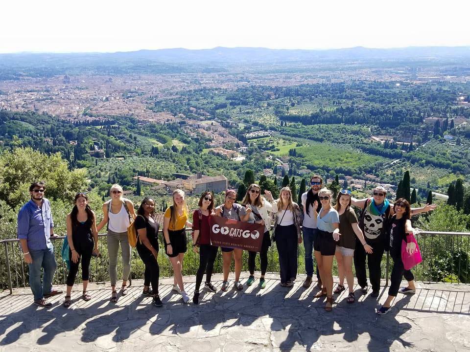 A group of TXST students pose for a photo on a cliff in Italy with a TXST sign.