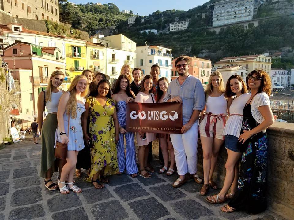 A group of TXST students pose for a photo outside a town in Italy with a TXST sign.