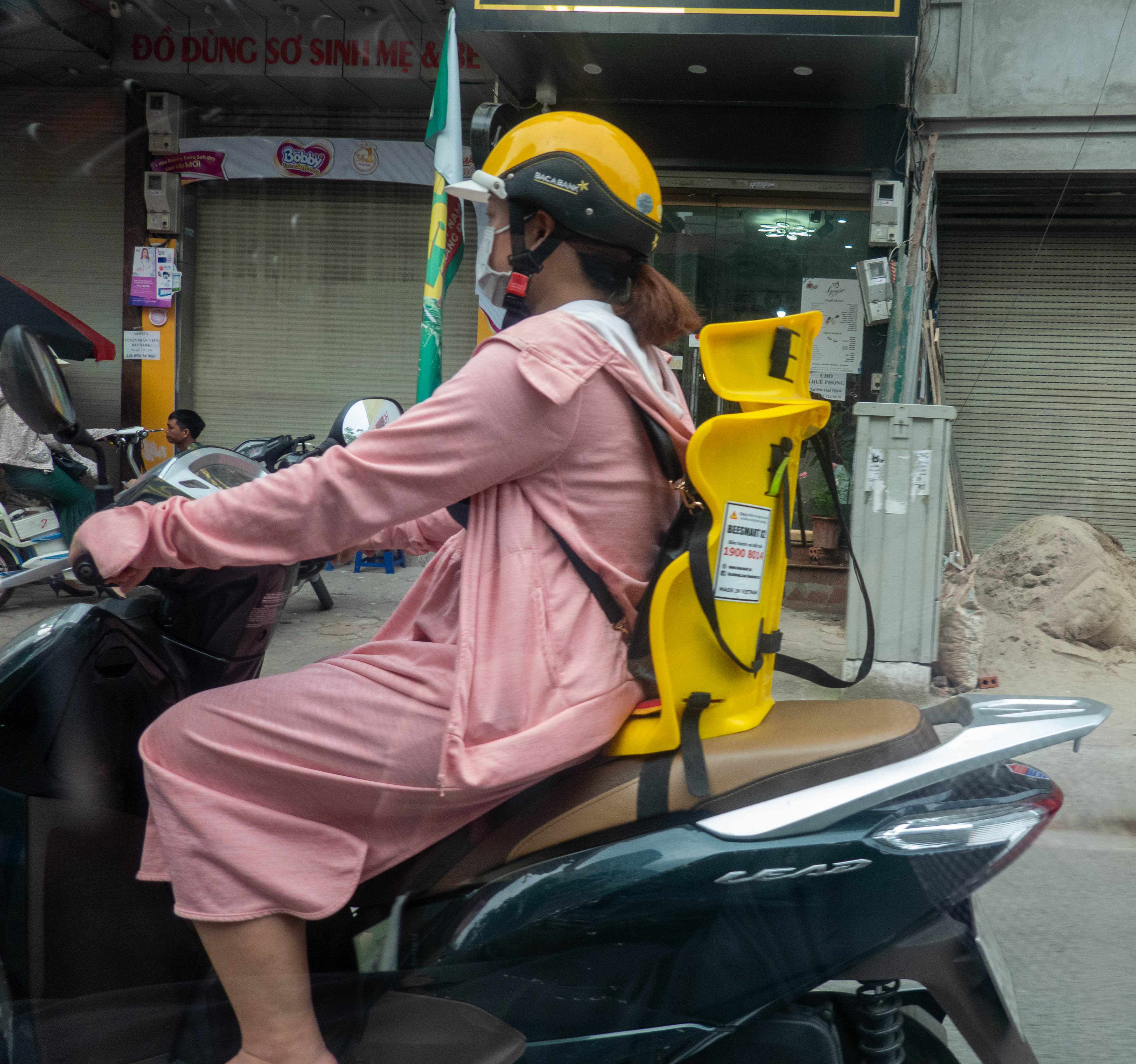 lady wearing yellow helmet on a motorbike. A yellow child holder is strapped to her back.