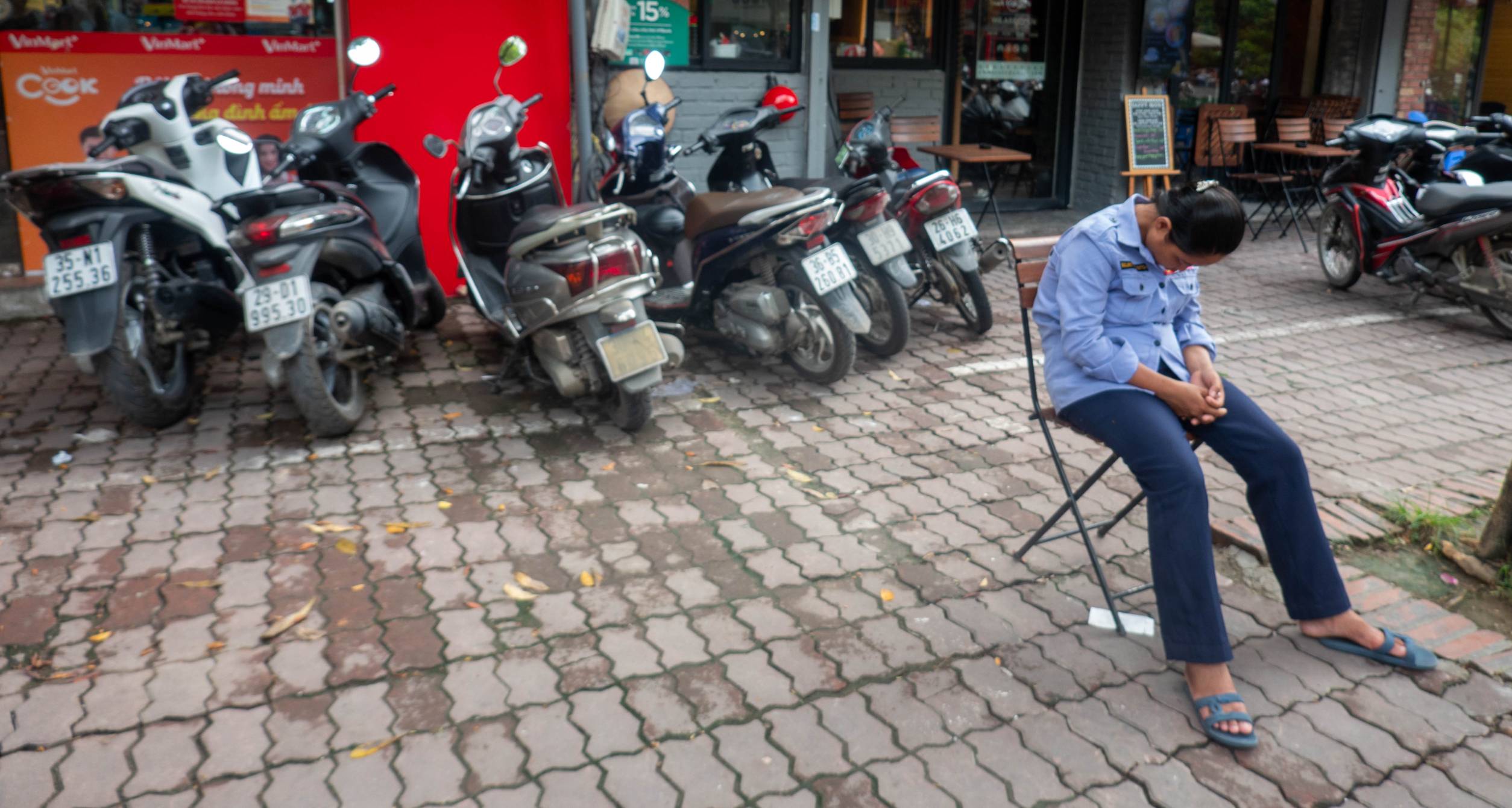 picture shows motorbikes parked in front of a business and in the foreground on the right is a women who is sitting in a chair who has fallen asleep.