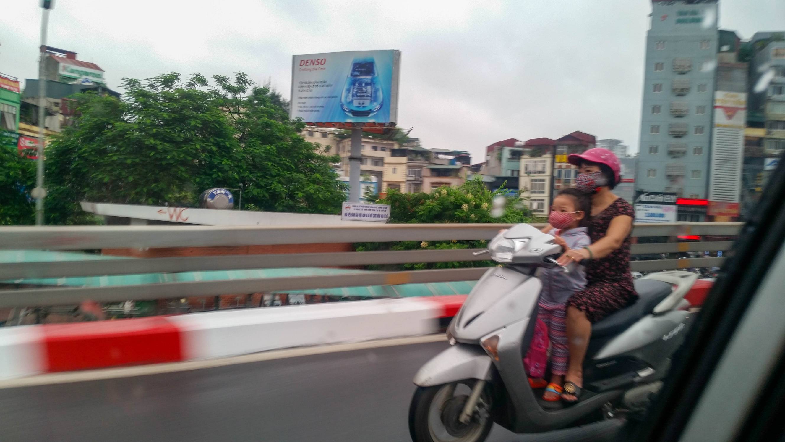 Toddler standing on the floorboard in front of the mother driving the motorbike. the child is holding on to the handle bars.