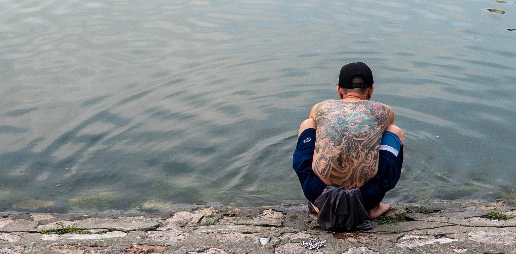 man squatting in front of lake with dragon tattoo on back