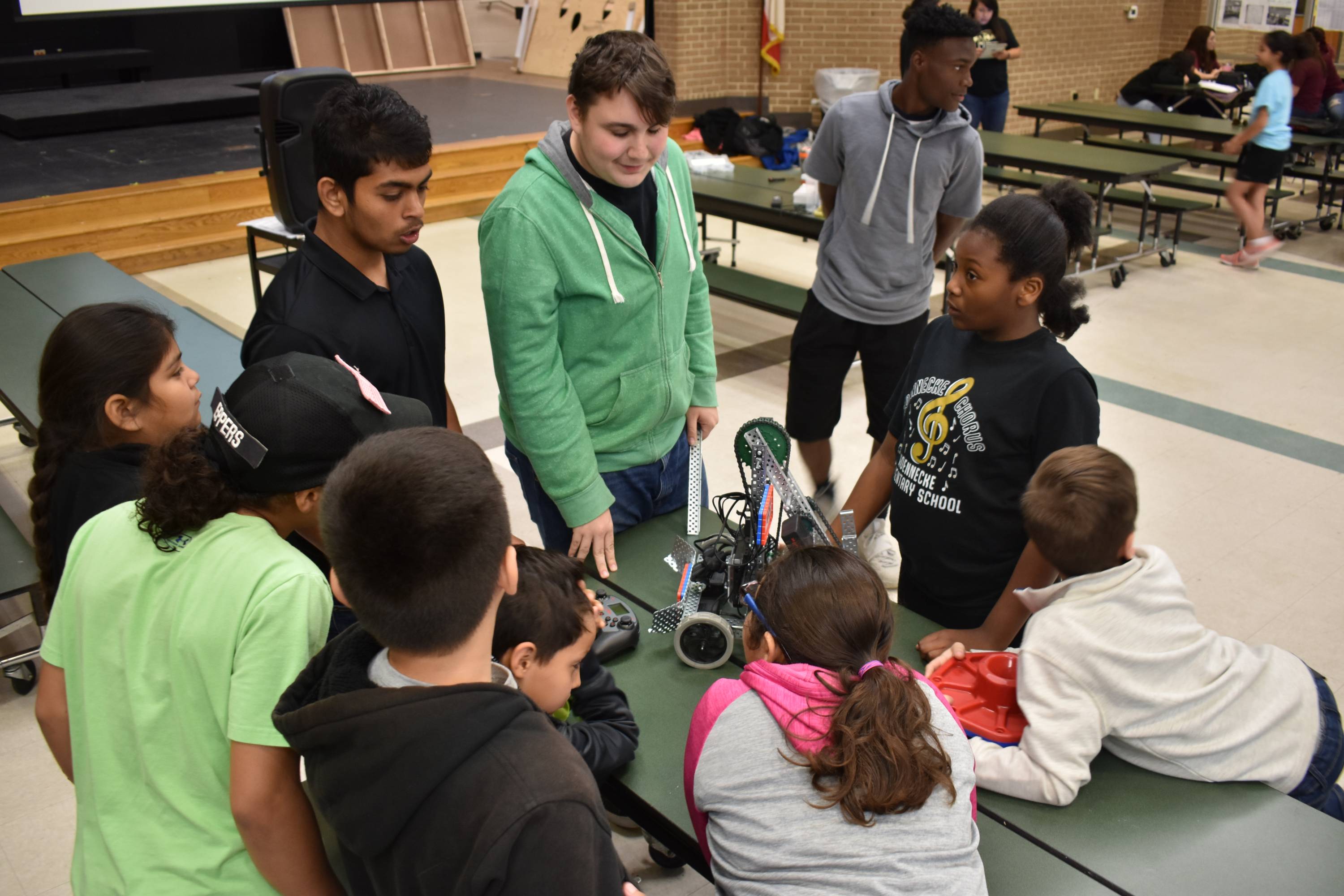 high school students show a group of younger students a robot that is positioned on a table in the middle of the group