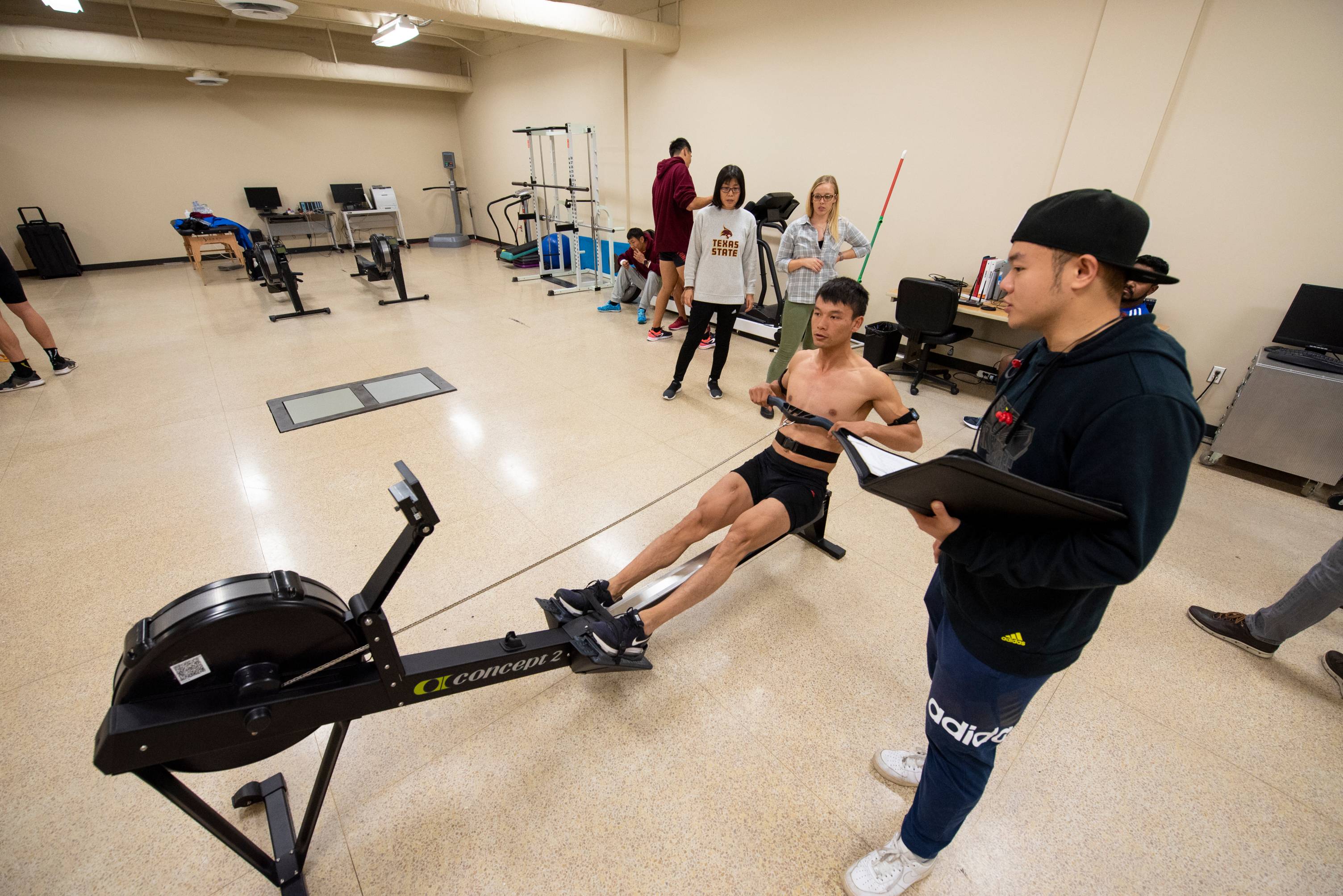 an member of the team uses a rowing machine while a graduate student records notes. Texas State faculty look on in the background.