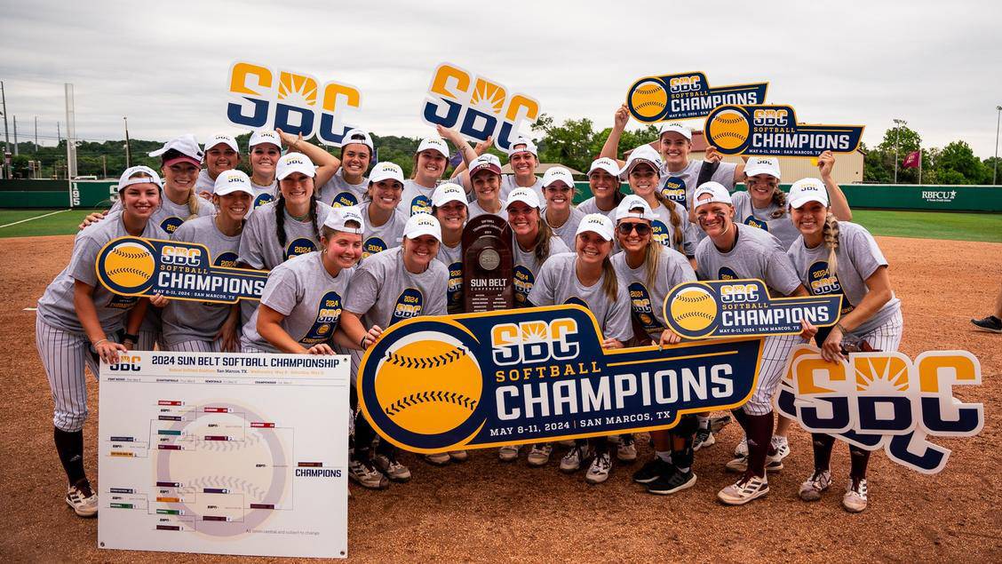 The TXST Softball team poses for a photo on the softball field with a Sun Belt Championship sign.