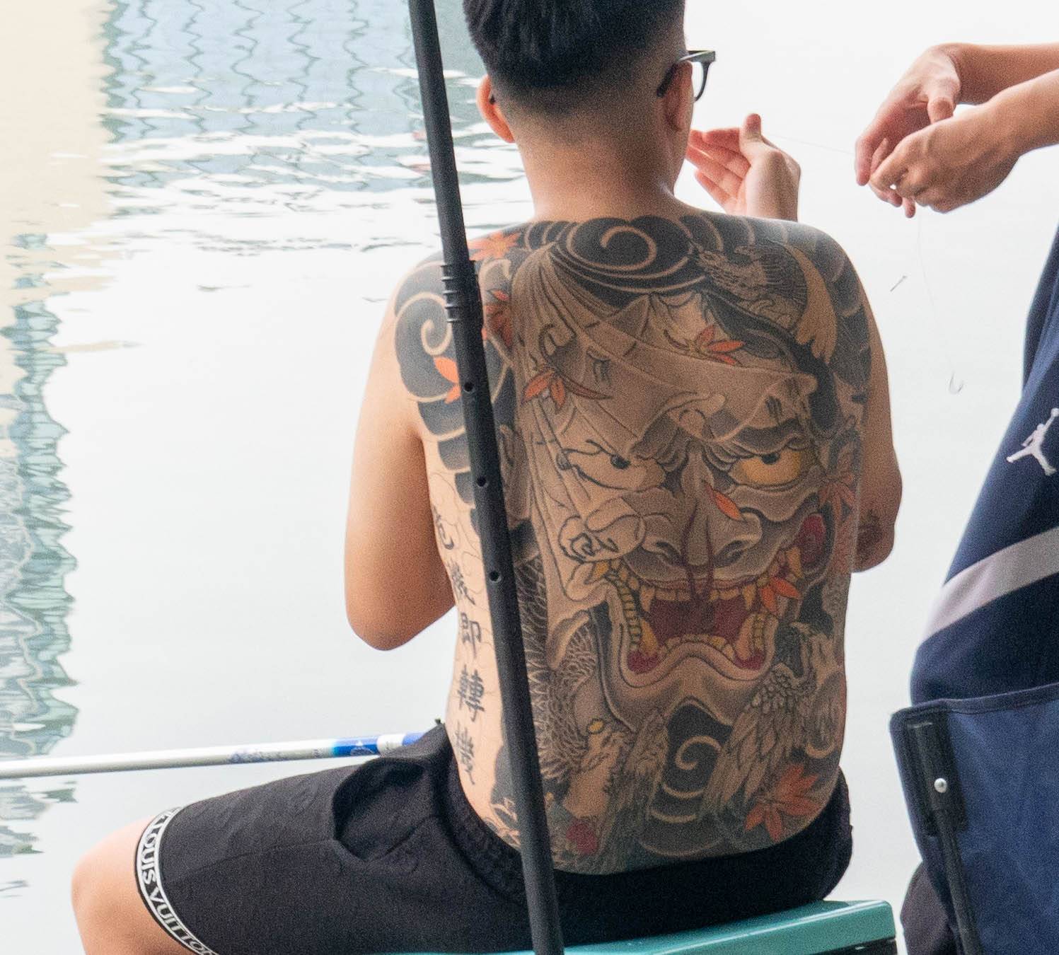 Picture of man in front of water looking directly away from you with a large tattoo of a dragon on his back. Picture taken from left side of man. dragon eyes appear to be looking right at you..