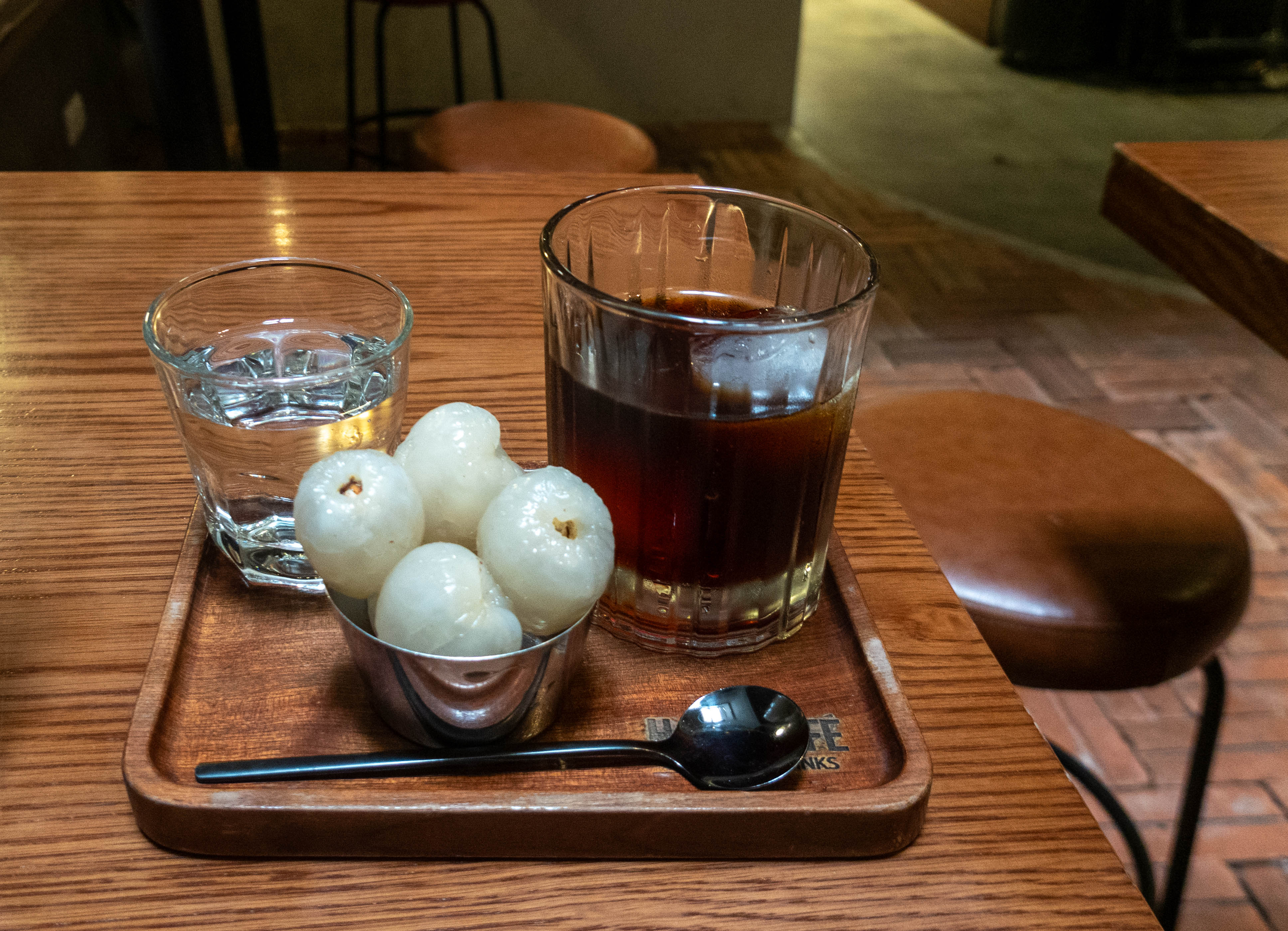 picture of a small glass of water (on left), a large glass of coffee with an ice cube (on right), and a small metal cup with four white transluscent oval shaped fruit (center) and there is a spoon in front of the metal cup. all are setting on top of a wooden tray on a wood table. there is a stool in the background on the right of the table.