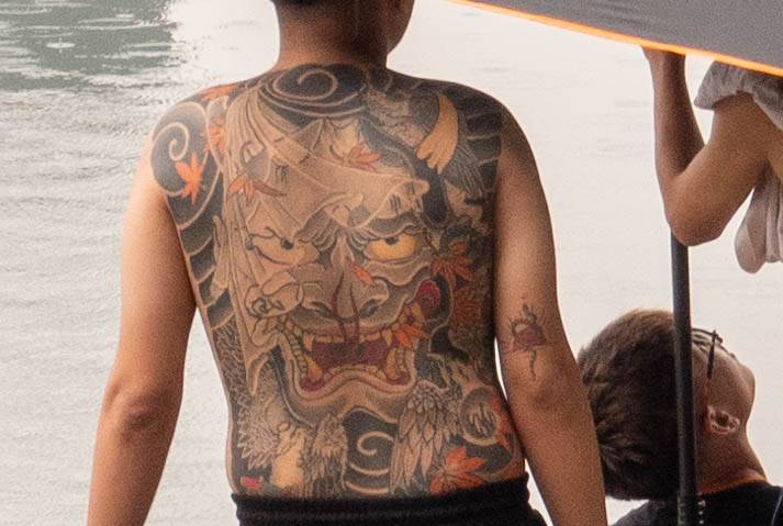 Picture of man in front of water looking directly away from you with a large tattoo of a dragon on his back -dragon eyes appear to be looking right at you..