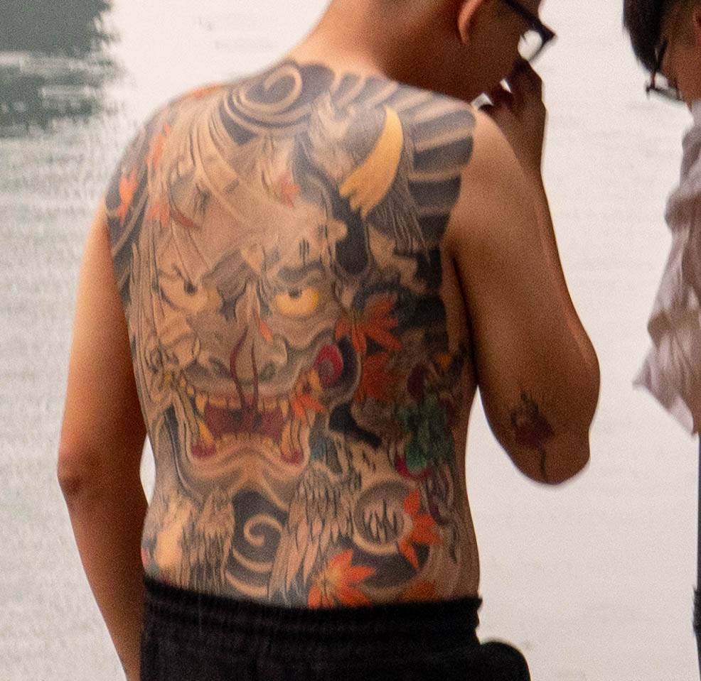 Picture of man in front of water looking away from you but turned to the right approximately 25 degrees, with a large tattoo of a dragon on his back -dragon eyes appear to be looking right at you..