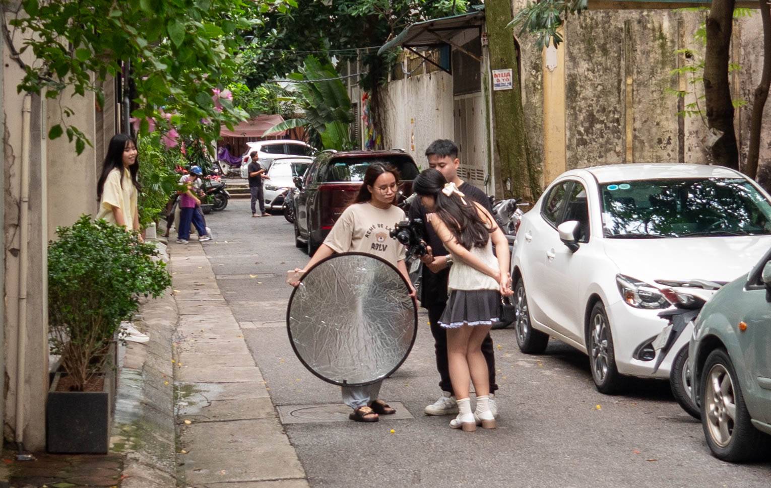 There is a woman with long black hair to the left of this picture looking at a group of three people looking at a camera.. one woman(on the left), a is holding a large round silver disc, a man is holding the small movie camera, and another woman is bending over looking at the camera. The scene is takend down a long alleyway with cars lining th right side.