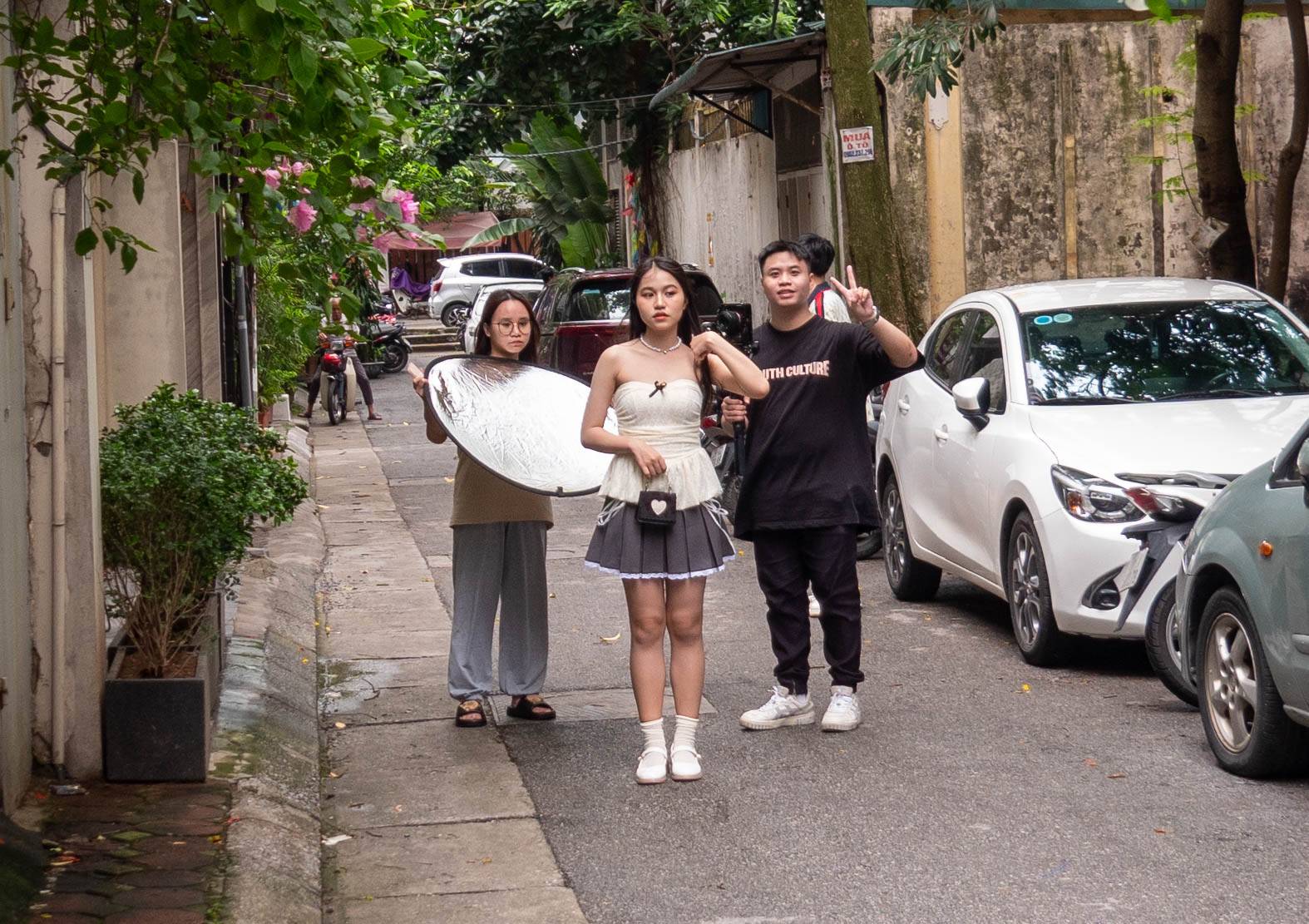 There is a woman with long black hair to the left is gone in this picture. The group of three are now looking at me as I take the picture. The man has his left hand up with his first two fingers making a "V". The scene is takend down a long alleyway with cars lining th right side.