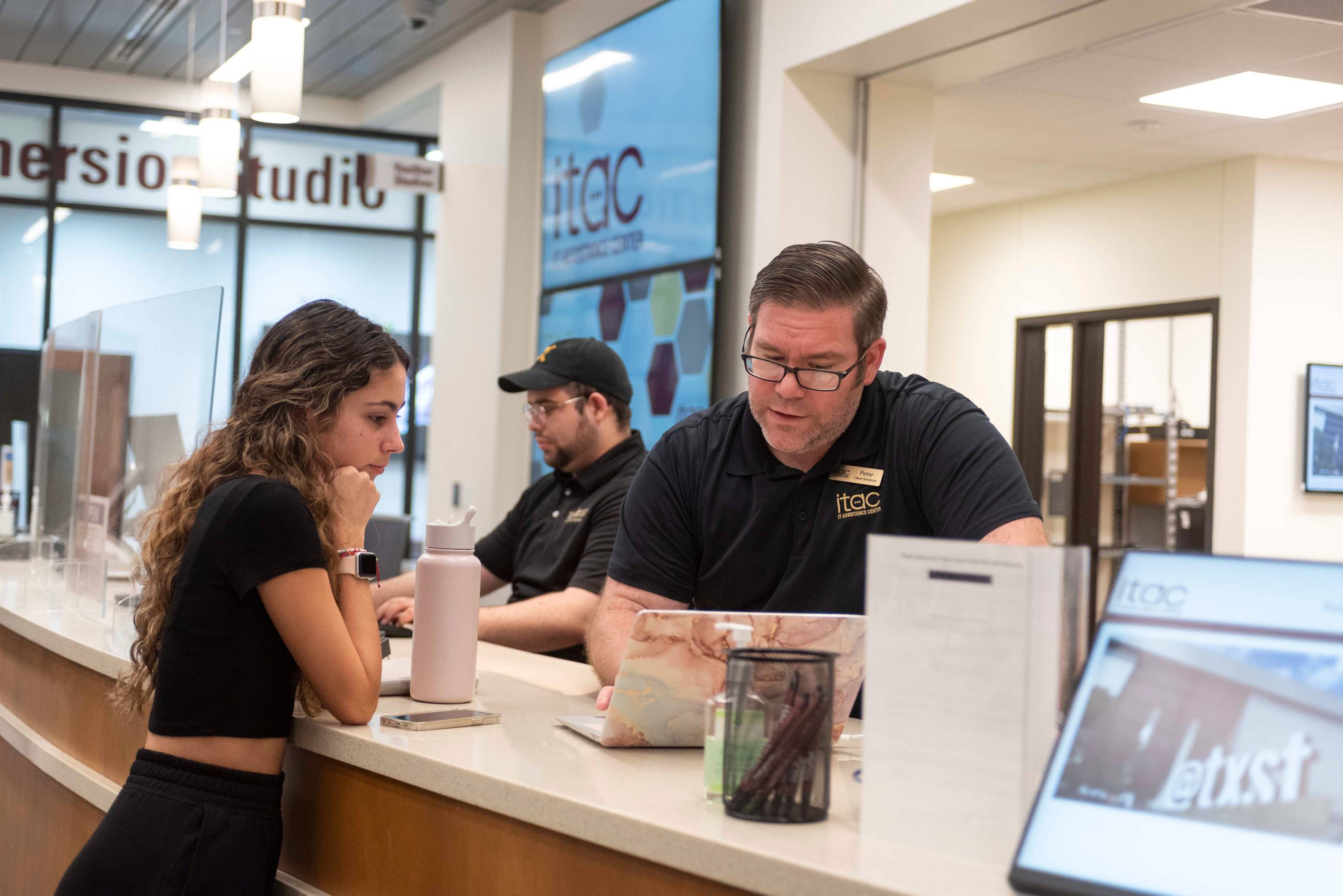 Man wearing a polo shirt and an IT Assistance Center badge points to something on the laptop of a girl learning over the counter.