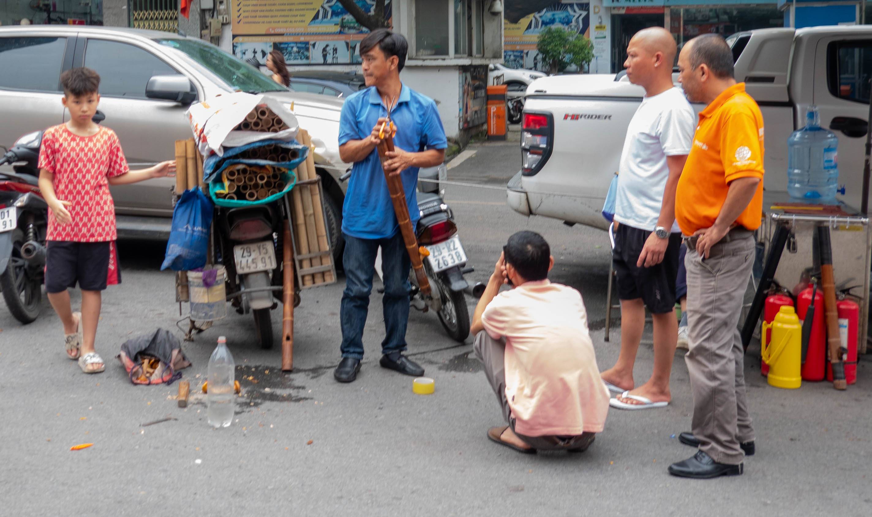 picture of four men and on young boy. Men ara looking at something off-camera to the left. Man in middle with blue shirt is holding two brown lengths of cut bamboo. Boy in red shirt is walking toward camera. There is a man squating down to left of man on right with yellow shirt.