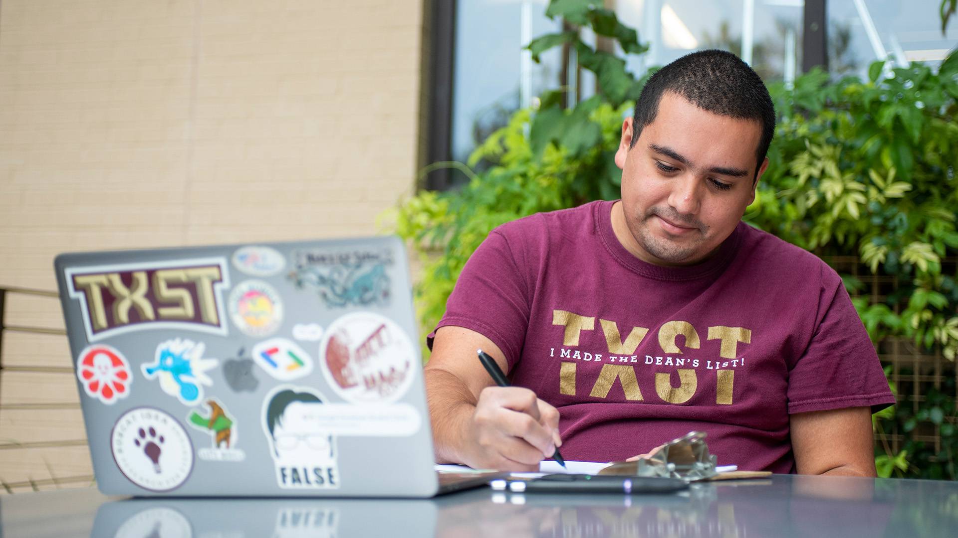 Student writing notes and studying with a laptop wearing TXST shirt that says "I made the dean's list"