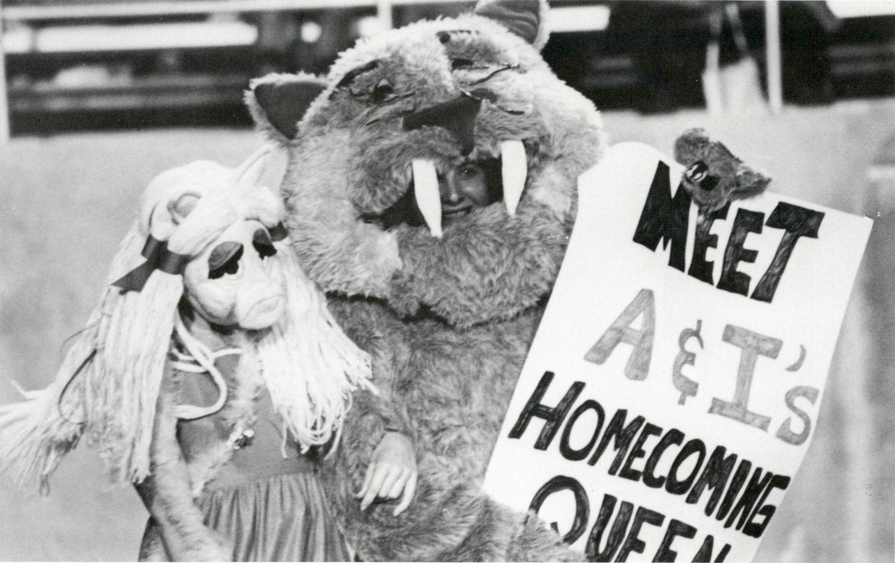 Black and white image of a woman in a bobcat mascot costume carrying a Miss Piggy muppet in one hand and a sign in the other that reads "Meet A & I's Homecoming Queen"