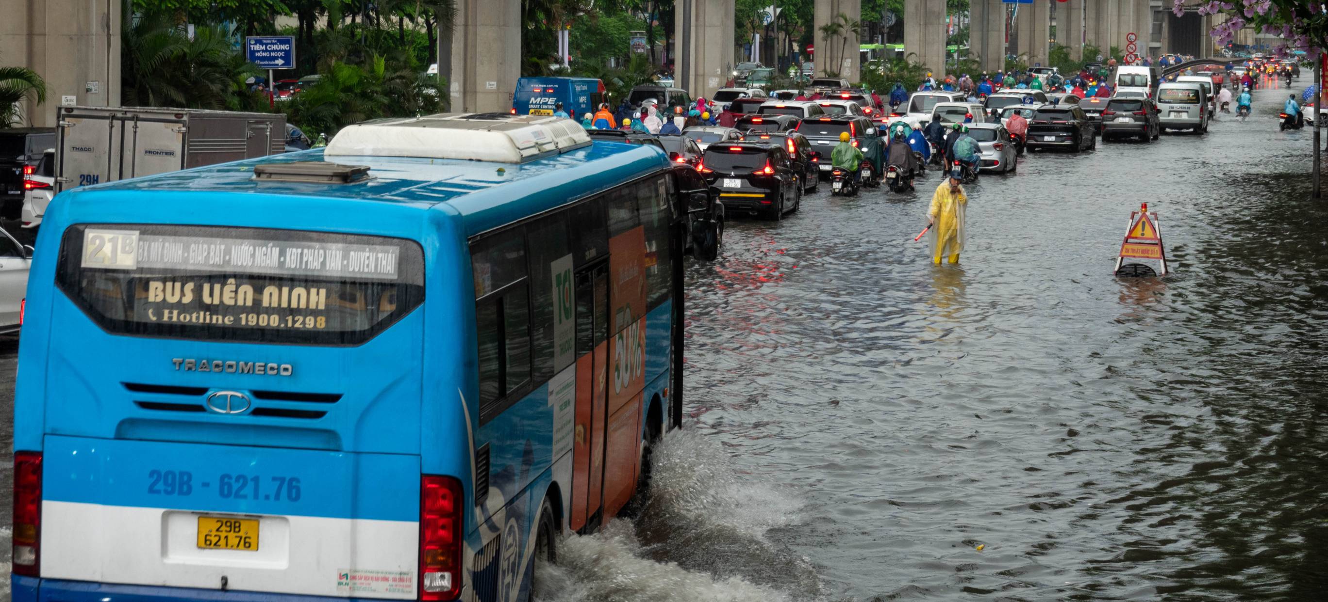 Traffic in deep water. Blue bus in foreground. Man in yellow standing in the water near a warning sign.