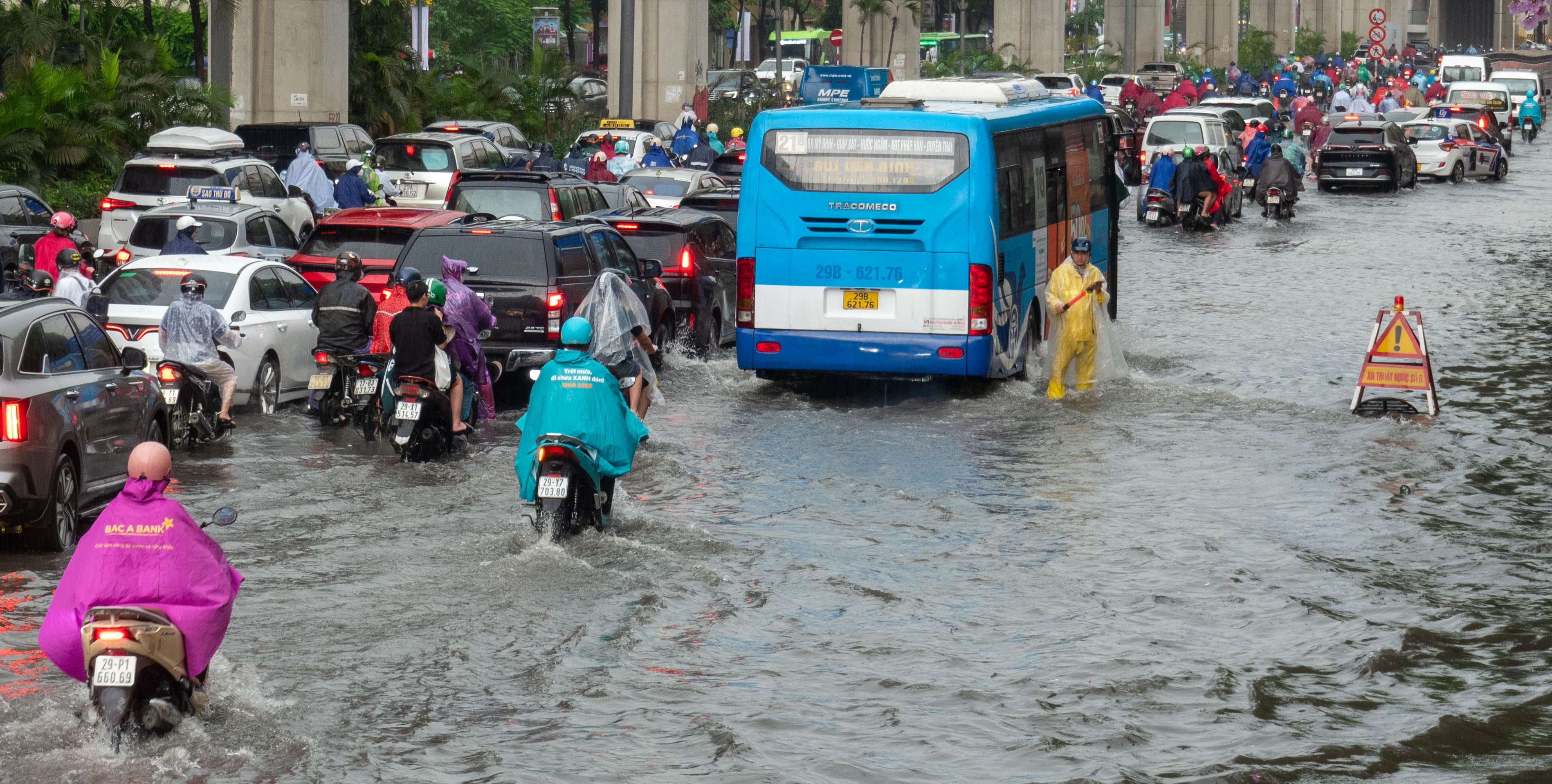 Traffic in deep water. Blue bus in foreground. Man in yellow standing in the water near a warning sign.