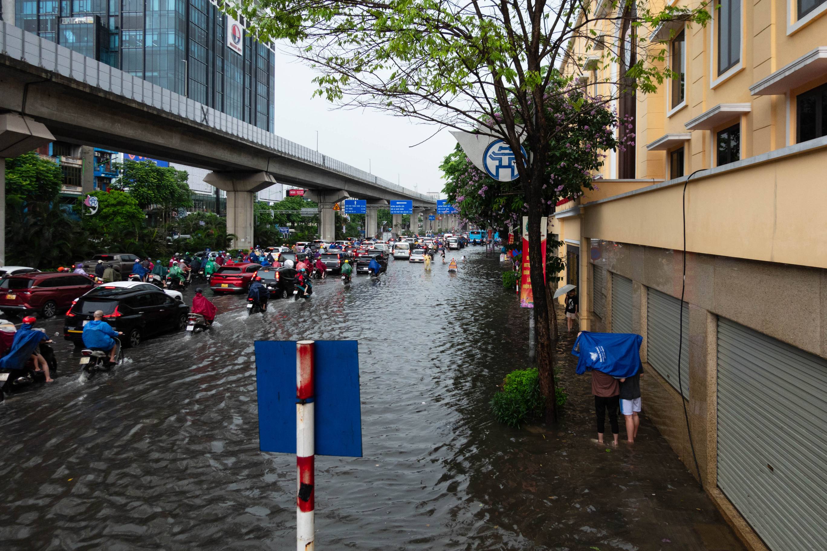 picture of flooded street with traffic and two people in the lower right of picture holding a blue piece of plastic over their head.